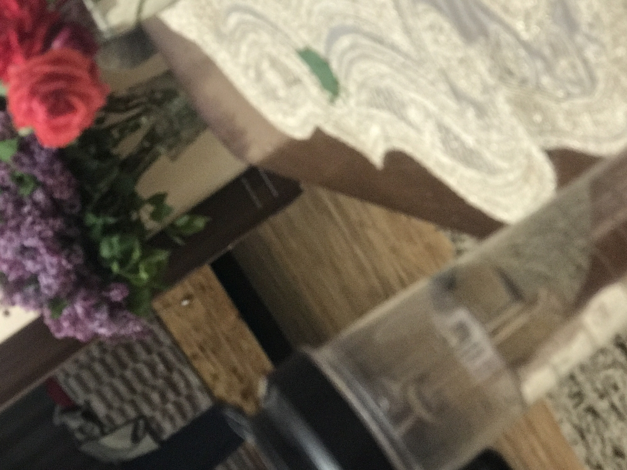 A vase of lilacs and roses sits on a table covered with a white lace tablecloth. A clear plastic container with a black lid is also on the table. The table is made of dark wood. The location is unknown and there is no evidence to suggest this is in Yerevan, Armenia.
