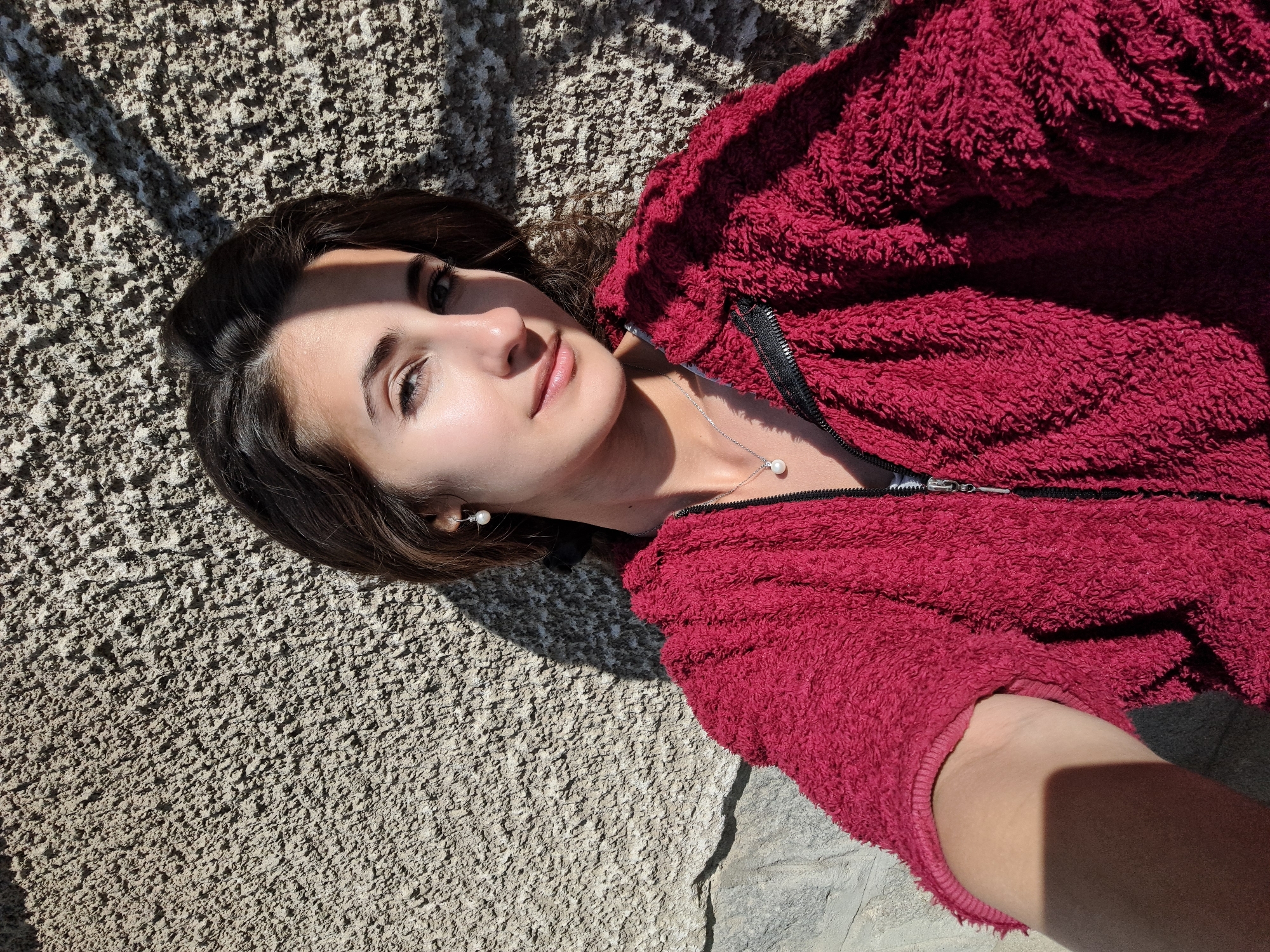 A young woman is depicted lying on a textured, light gray or beige ground surface, possibly concrete or a paved area. She is looking upwards towards the camera with a gentle, relaxed smile. Her dark hair is splayed around her head. She wears a dark red, fuzzy, long-sleeved jacket with a visible black zipper running down the front. A delicate silver necklace with a single white pearl pendant rests on her chest, and she has matching pearl stud earrings.
The scene is brightly lit by direct sunlight, casting sharp, distinct shadows on the ground and partially across her face, suggesting a clear, sunny day, likely mid-morning or early afternoon. The texture of the ground is prominent, appearing rough and granular. One crop hints at a slightly different stone texture beneath her arm, indicating a varied outdoor paving. The woman's pose and expression convey a sense of calm and contentment. There is no visible text in the image.