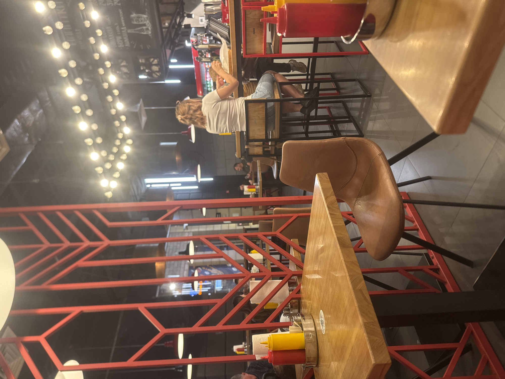 The image depicts the interior of a dimly lit restaurant, possibly a fast-casual establishment with a modern industrial design, featuring red metal accents and string lights. In the foreground, a wooden table with a matte finish is visible, set with a metal caddy containing red and yellow squeeze bottles, likely ketchup and mustard, along with napkins. A brown leather-style chair with black metal legs is positioned near this table.
Further into the restaurant, a woman with blonde hair pulled into a bun is seated at a bar, facing away from the camera. She is wearing a white t-shirt and denim shorts and appears to be eating or examining food. Behind the bar, various kitchen equipment and a red refrigerator are visible. Other patrons are seated at tables in the background, and the overall atmosphere suggests a casual dining environment. The restaurant's decor includes a prominent red geometric partition in the midground, separating different seating areas. The floor is dark and appears to be tiled, reflecting the ambient light.
The location is identified as Sovkhoza imeni Lenina, Russia.