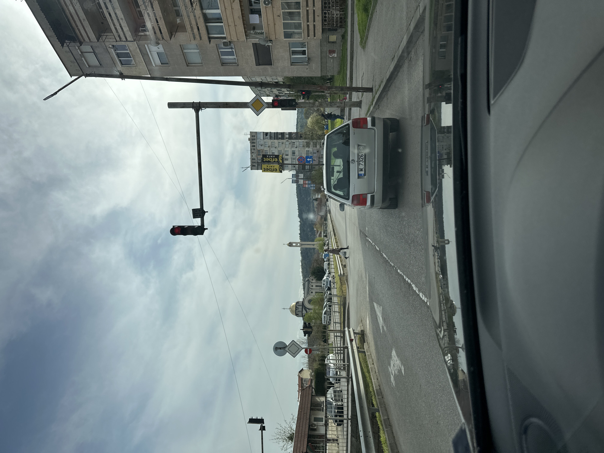 The image depicts a street scene in Montana, Bulgaria, viewed from inside a car. The car is stopped at a red traffic light. Another silver car is ahead, displaying a license plate number "M 7326 PC". The buildings along the street consist of residential apartments and commercial establishments. In the distance, a church with a golden dome and a tall clock tower is visible. Traffic signs are present on the roadside. The sky is cloudy, suggesting an overcast day.