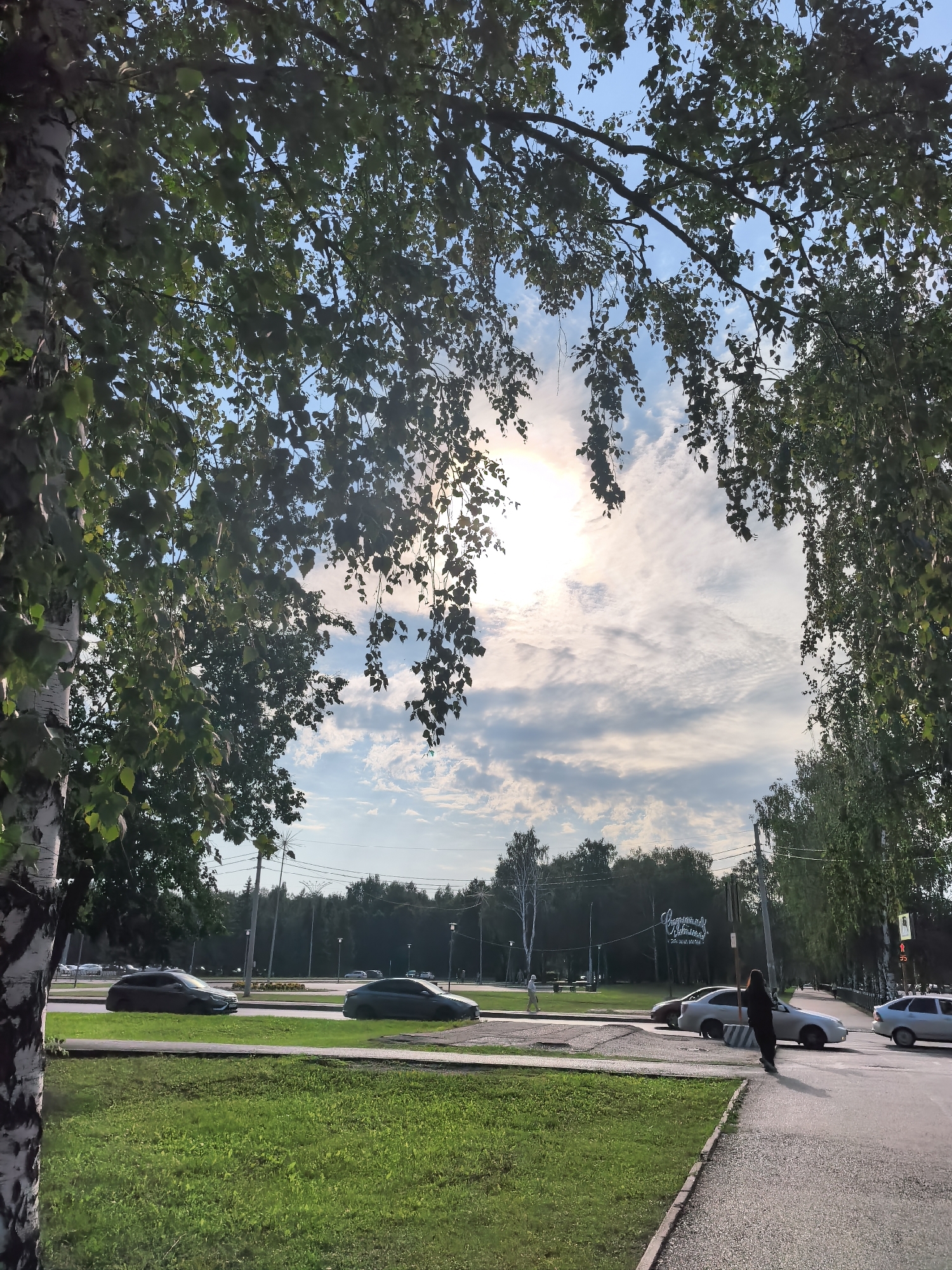The image captures a street scene in Sterlitamak, Russia, likely during the daytime with the sun partially obscured by clouds. In the foreground, a sidewalk runs along a grassy median strip. A person dressed in dark clothing is walking on the sidewalk. Beyond the median, a multi-lane road has several cars, including a dark sedan on the left, a light-colored sedan in the middle, and a silver sedan on the right. A sign with Cyrillic lettering is visible in the background, situated among trees. The trees, with their green foliage, frame the upper portion of the image, with a prominent birch tree on the left. The sky is a mixture of blue and white clouds, with the sun creating a bright glare. The overall impression is a calm, everyday urban environment.
