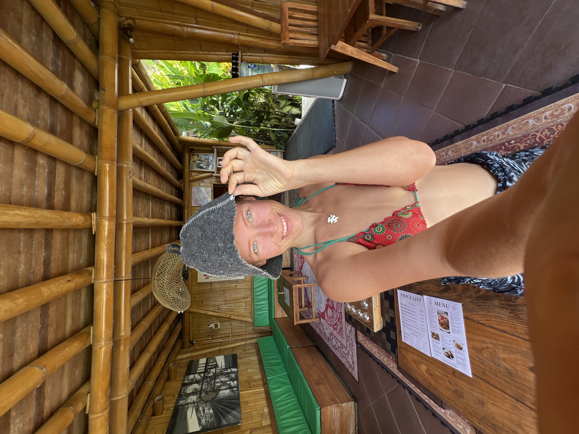The image shows a woman taking a selfie in a bamboo structure in Ubud District, Indonesia. The structure has bamboo walls and supports. Inside, there is seating with green cushions, a bamboo fan, and framed artwork on the wall. A table has a price list and menu. The woman is wearing a red patterned bikini top, a gray hat, and a necklace. Outside, greenery can be seen. The floor is tiled.