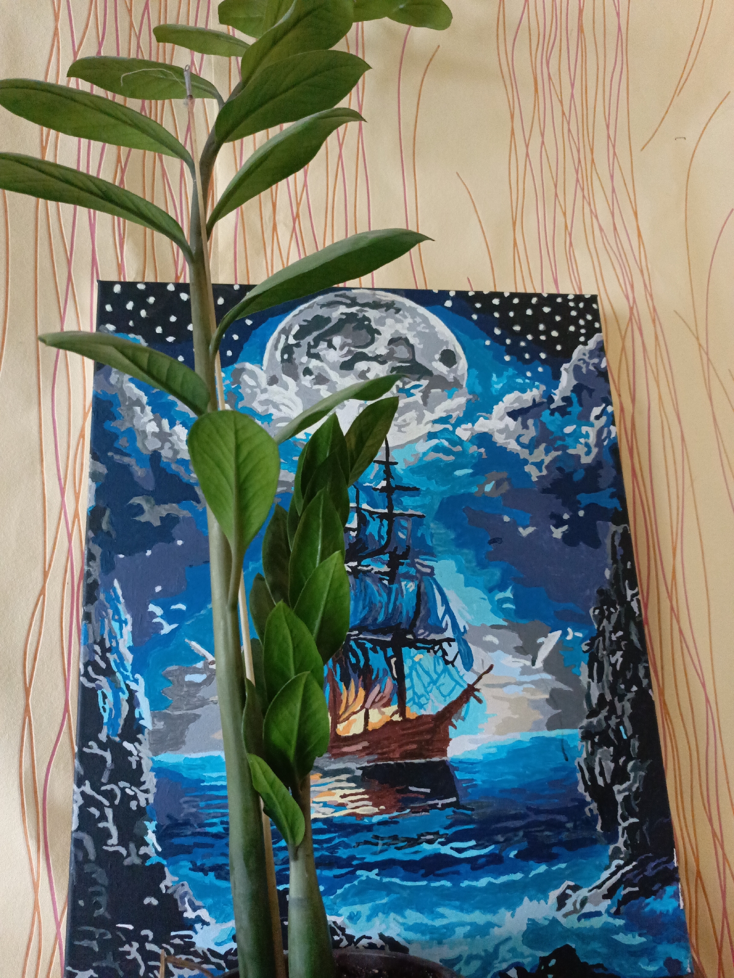 The image shows a plant in the foreground with a painting behind it. The painting depicts a sailing ship on the ocean at night, with a large moon in the background surrounded by clouds and stars. The painting has a border on the left and bottom edges. The background is a wall with vertical wavy stripes. The image was taken indoors and no people are visible. There is no evidence in the image to indicate it was taken in Zalesovo, Russia.