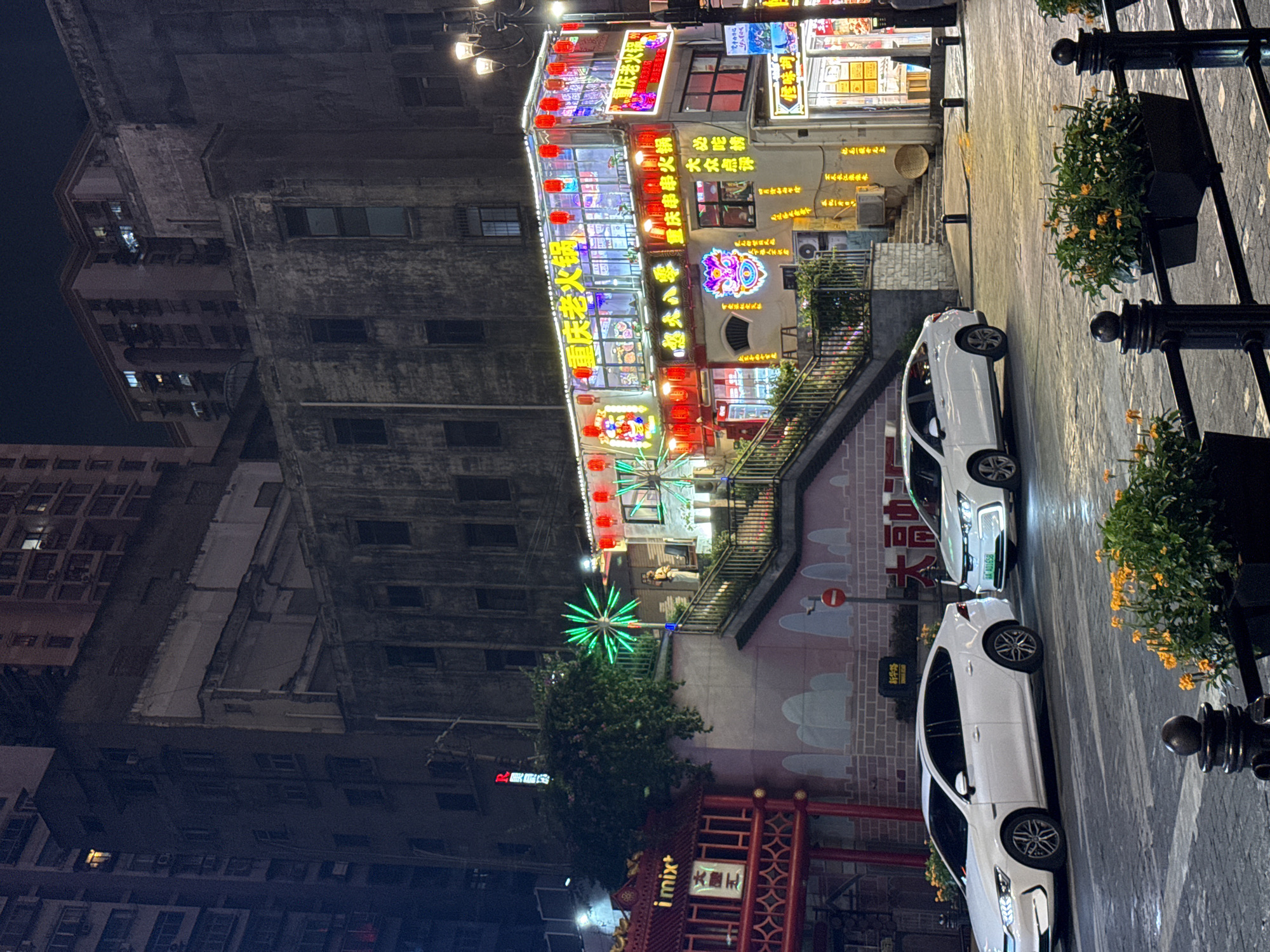 The image depicts a street scene at night in Yuzhong District, China, likely a commercial or entertainment area. A multi-story building with illuminated signage and numerous red lanterns is prominently featured. The signage is in Chinese characters, with one prominent sign reading "重庆老火锅" (Chongqing Hot Pot). The building appears to be a restaurant or a cluster of shops.
In the foreground, two white sedans are parked on the street. The car on the left has its headlights on, illuminating the road. The car on the right is also illuminated, suggesting it is either parked with lights on or has its daytime running lights active. The street is paved with cobblestones or a similar textured surface.
To the left of the illuminated building, there's a red structure with traditional Chinese architectural elements and signage, one reading "i mix+". A staircase with metal railings leads up to the illuminated building. Green neon lights shaped like a starburst are visible near the staircase.
The background shows the dark facades of older, taller buildings, some with lit windows, indicating residential or office spaces. The overall atmosphere is vibrant due to the colorful lighting of the restaurant, contrasting with the darker, more subdued buildings in the background and the nighttime setting. The street also has planters with orange flowers and black decorative railings in the foreground.