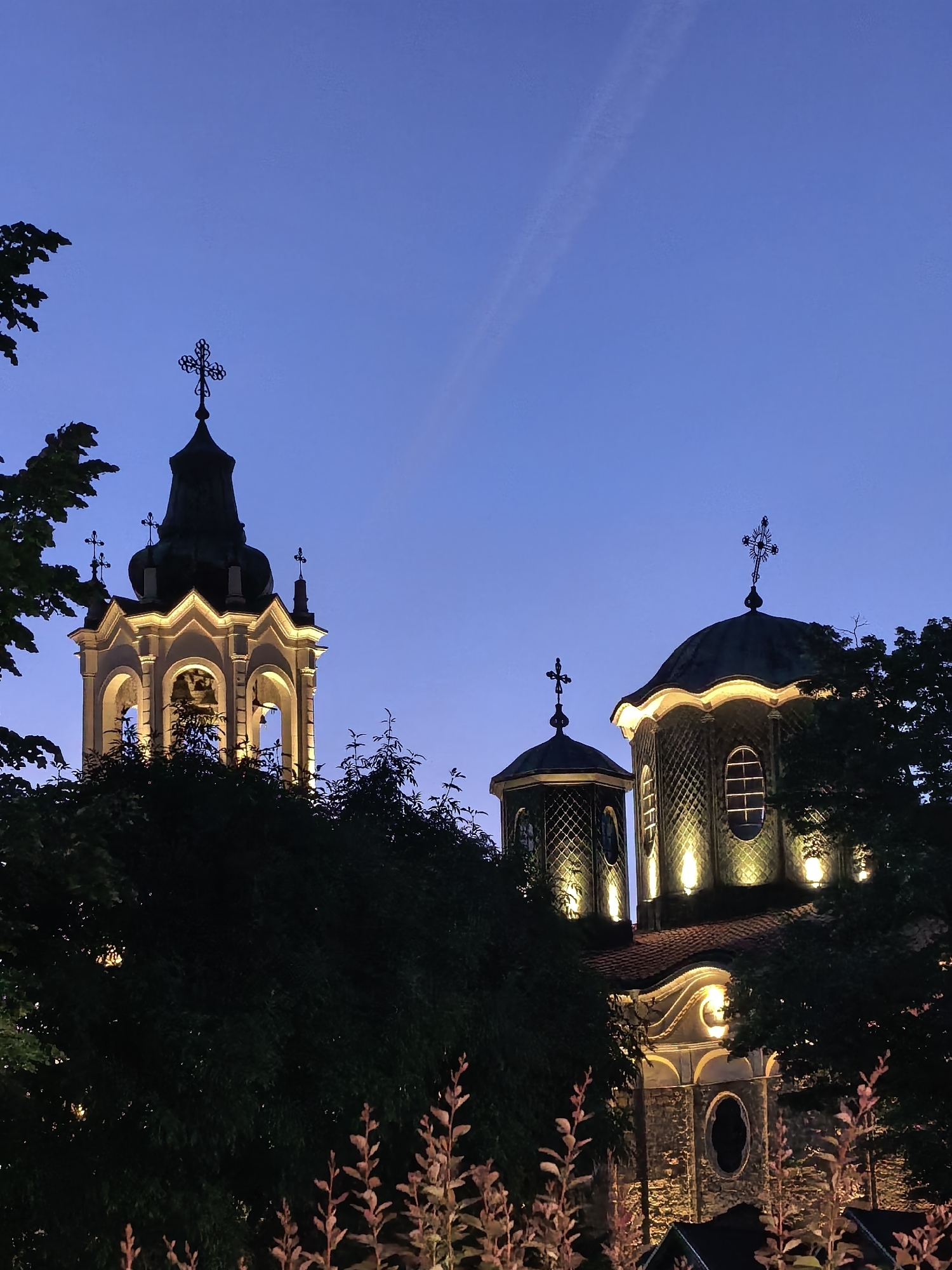 The image captures the illuminated architecture of the Holy Trinity Church in Svishtov, Bulgaria, against a twilight sky. A church bell tower stands on the left, and the church domes on the right are partially obscured by trees. A faint contrail is visible in the sky above the church. Vegetation is present in the foreground, and the buildings are illuminated.