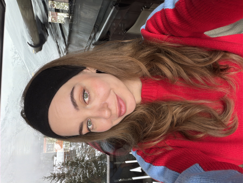 A young adult female is depicted in a close-up, chest-up shot, gazing directly at the camera with a slight smile. She has long, wavy, light brown hair worn down, partially covered by a black fabric headband. Her facial features include light green eyes, defined eyebrows, and light-toned makeup. She wears a red knitted sweater featuring light blue vertical stripes on the upper arms and shoulders. The subject is positioned indoors or on a sheltered balcony, with a winter outdoor scene visible through a reflective glass partition or window in the background. The background reveals snow-covered evergreen trees and distant snow-dusted terrain under an overcast sky, characteristic of an alpine environment. Elements of a building, including a dark railing structure and a snow-laden roof with a dark pipe or fixture, are also present in the background. The scene is located in the city of Les Allues, France.