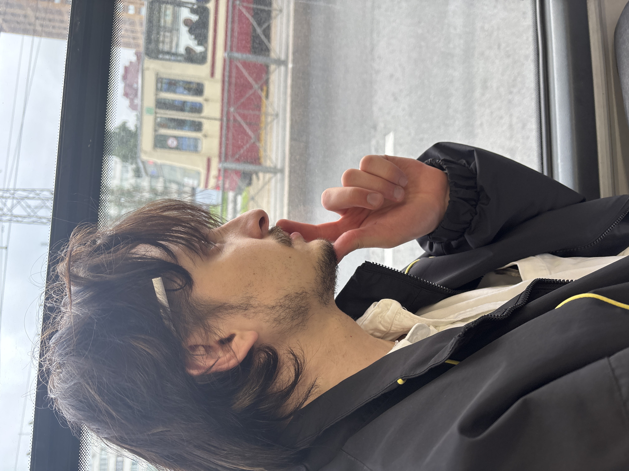 A young man with dark, medium-length hair and stubble is sitting inside a tram in Saint Petersburg, Russia, looking out the window. He is wearing a black jacket over a white shirt. His right hand is raised to his chin, with his index finger touching his lips, suggesting a contemplative or thoughtful mood. Outside the tram, a yellow and red tram with passengers can be seen on the tracks, with buildings and a cloudy sky in the background. The lighting is overcast, indicating a possibly rainy or gloomy day.