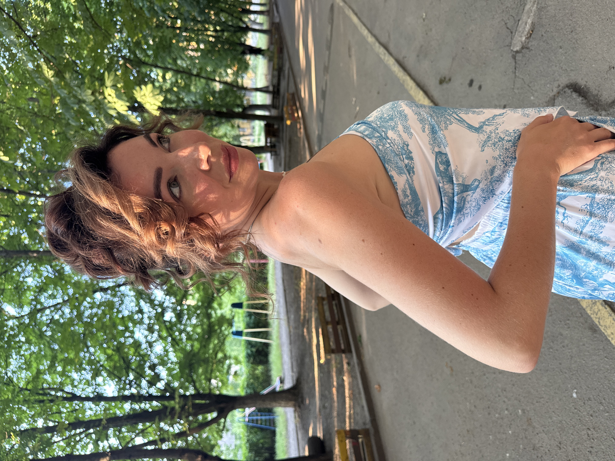 The image shows a young woman with auburn hair, who is looking towards the camera with a slight smile. She is wearing a white and blue patterned dress. The woman is outdoors, with trees and greenery in the background. Sunlight is dappling through the leaves, casting shadows on the ground and on her face. The setting appears to be a park or a recreational area, possibly with a playground visible in the distance. The lighting suggests it is daytime, likely late afternoon due to the long shadows. The overall mood is relaxed and pleasant. No visible text is present in the image. The location is identified as Kryvyi Rih, Ukraine.