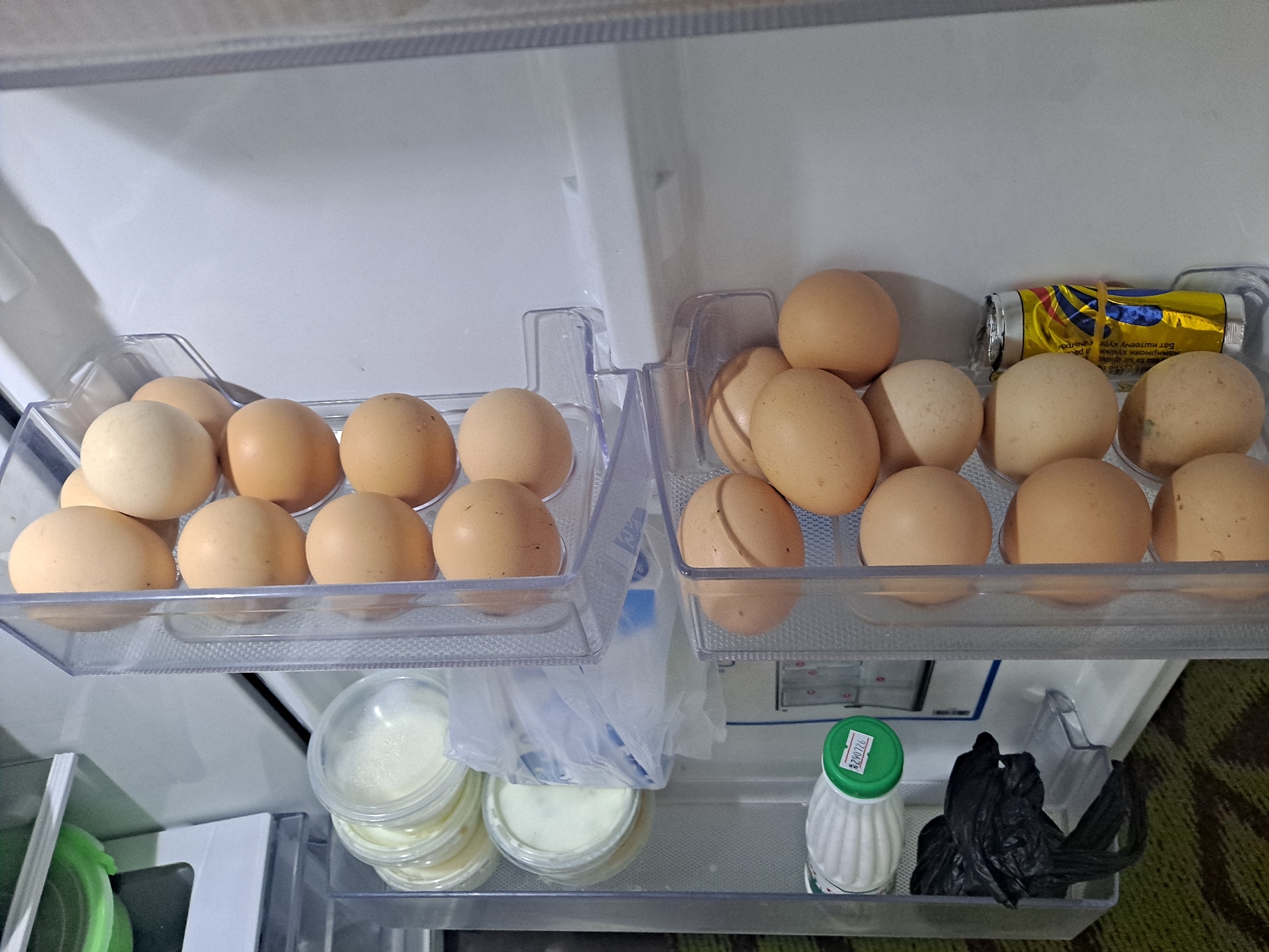 The image shows the interior of a refrigerator. On the top shelf, there are two clear plastic egg holders filled with brown and white eggs. On the right side of the top shelf, there is a silver and yellow foil package secured with a rubber band. On the bottom shelf, there are several small plastic containers filled with a white substance, a green-capped bottle with a white liquid inside, and a crumpled black plastic bag. The refrigerator appears to be in a domestic setting.