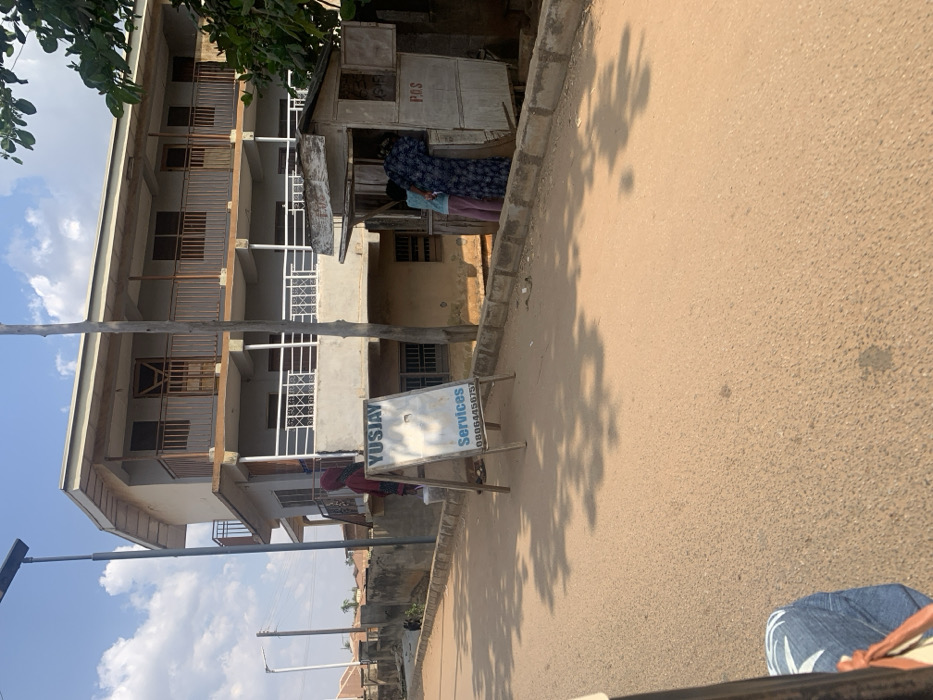 The image depicts a sunlit street scene in Okengwe, Nigeria, when viewed upright. On the left side, a multi-story building with several balconies, some featuring white railings and others brown wooden ones, dominates the mid-ground. A tall utility pole stands vertically in the left foreground, while a more distant pole with a street light, possibly solar-powered, is visible further down the street. Green foliage from a tree is seen at the top left, casting prominent shadows onto the road.
To the right, a small, makeshift wooden shack or kiosk is present. Inside, a person wearing a dark, patterned garment is partially visible. The letters "P.O.S" are painted on the side of this structure, indicating it likely offers point-of-sale or agency banking services. Another person in reddish attire can be faintly seen further down the street near the multi-story building.
The foreground and mid-ground are occupied by a paved road, likely asphalt, which extends into the distance. Distinct, relatively short shadows of trees are cast across the road, suggesting a sunny day during midday or early afternoon. An A-frame sign stands on the roadside in the mid-ground, clearly displaying "YUSJAY Services" and a phone number: "08064450757". The bottom right corner of the image shows a partial view of a person's legs and shoes, indicating the photograph was taken from a low vantage point, possibly from a vehicle or by someone walking. The overall scene conveys a calm, everyday atmosphere.