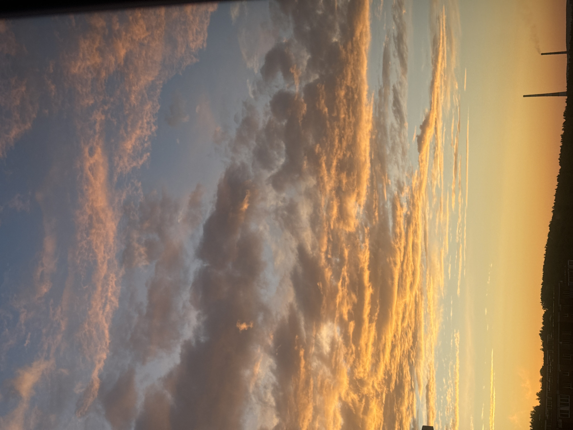 The image depicts a sunset sky over the city of Markova, Russia. Wispy clouds, tinged with orange and pink hues, stretch across a gradient of blue to yellow sky. In the foreground, a dark silhouetted treeline forms the horizon, with a portion of buildings visible beneath it. Two tall, slender smokestacks rise from the landscape on the right side of the image, with one emitting a faint plume of smoke. The overall scene is serene and atmospheric, characteristic of twilight in a temperate climate.