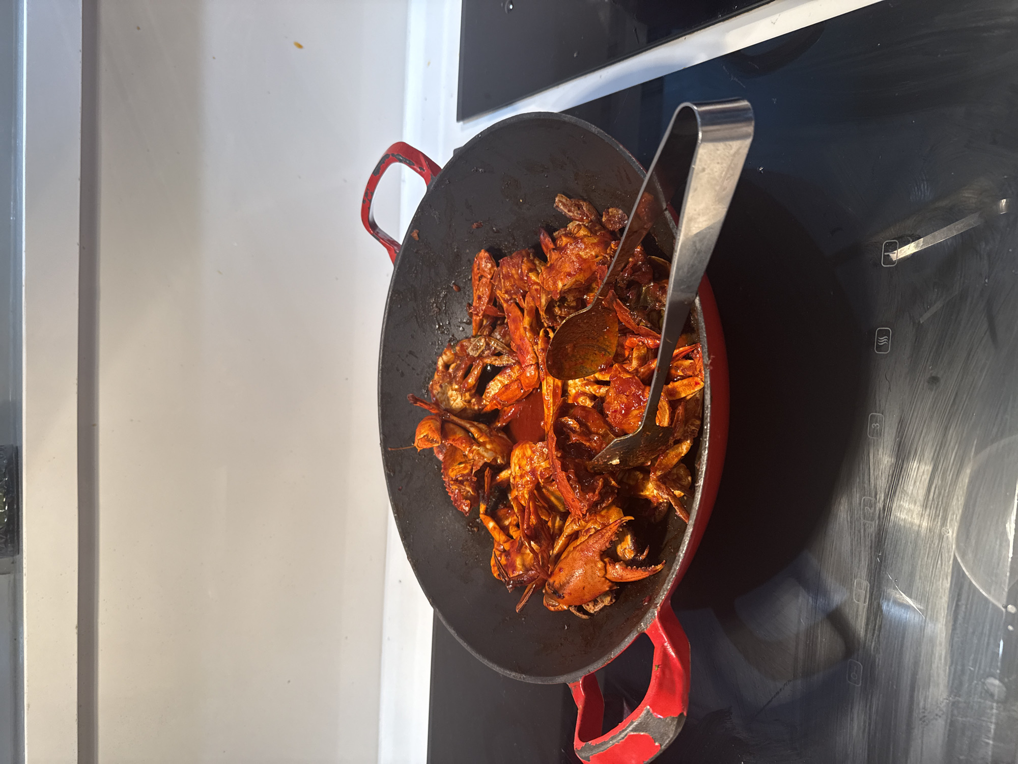 A close-up, eye-level shot shows a wok filled with spicy, red-sauced crabs on a black induction stovetop. The wok has red handles and appears to be made of cast iron. A pair of stainless steel tongs is resting on top of the crabs, with its handle pointing towards the right. The background is a blurred white surface and a white countertop. The image was taken in Nha Trang Ward, Vietnam.