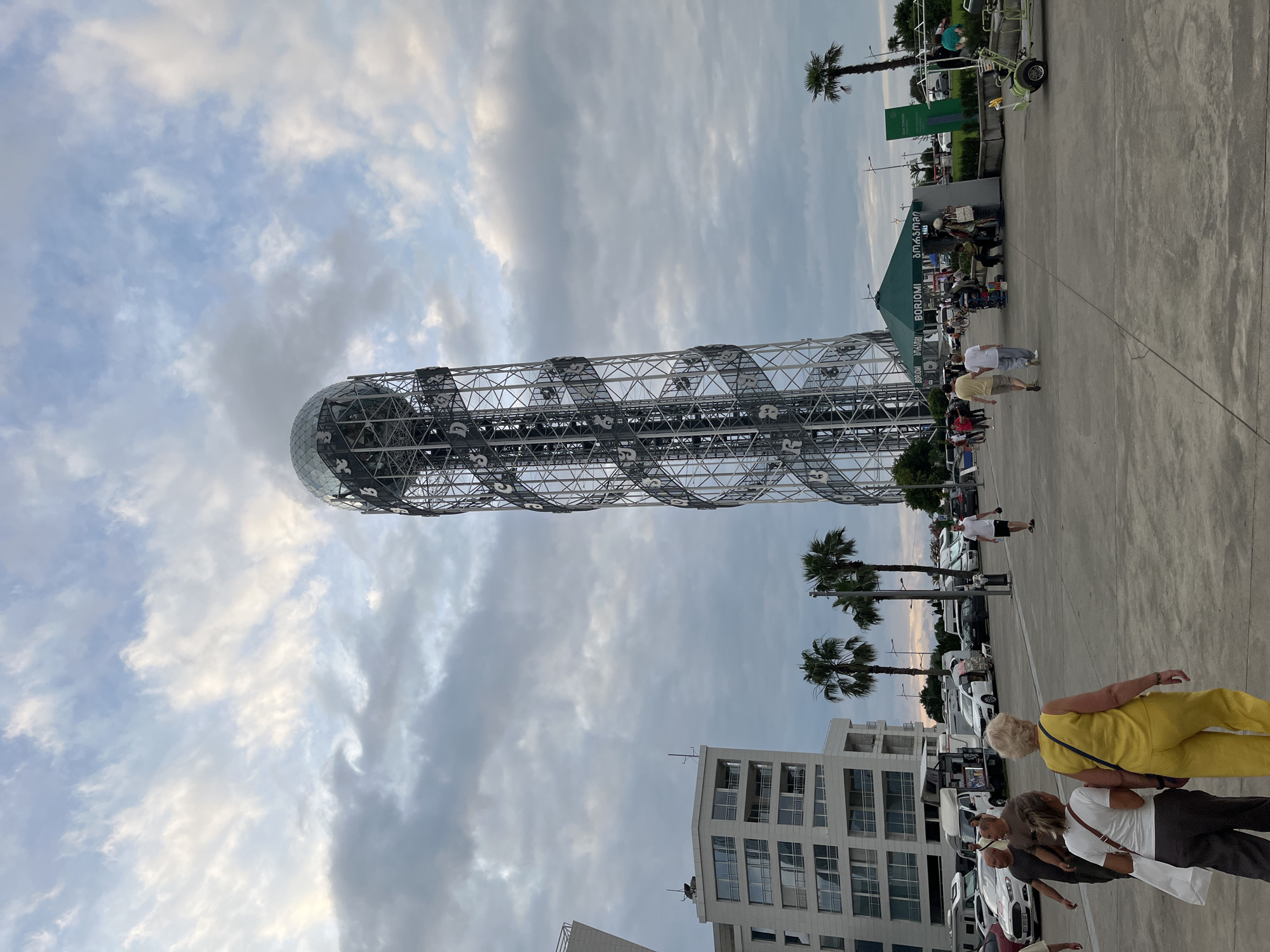 The image depicts an outdoor scene in Batumi, Georgia, with a prominent modern tower structure dominating the left side. The tower features a spherical top and a lattice-like metallic structure spiraling around its height. The ground is paved, suggesting a public area or promenade. Several people are walking on the paved surface, some individually, others in small groups. Their attire appears casual, consistent with daytime activity. In the background, palm trees add to the subtropical feel, and cars are parked in a lot, indicating transportation access. A multi-story building with numerous windows is visible towards the lower right, suggesting an urban environment. The sky is partly cloudy, with patches of blue and diffused sunlight, implying it might be late afternoon or early evening. Visible text includes "BORJOMI" on a green awning and also on signage attached to the green structure, possibly indicating a beverage brand or vendor. The overall impression is a leisurely day in a seaside city, with tourists or locals enjoying the area.