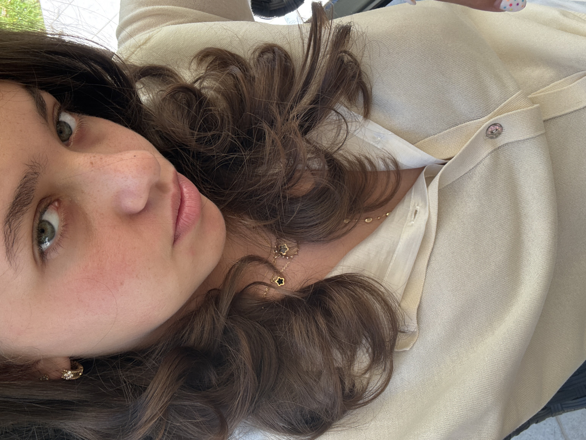 A close-up, high-angle shot shows the upper body of a young woman with brown hair, green eyes, and fair skin. She is wearing a cream-colored knit cardigan over a lighter cream-colored collared shirt. Her hair is styled in loose waves. A gold necklace with a black clover charm hangs around her neck. She is also wearing gold hoop earrings. The background is blurred and out of focus, but appears to be outdoors. The image was taken in Sofia, Bulgaria.