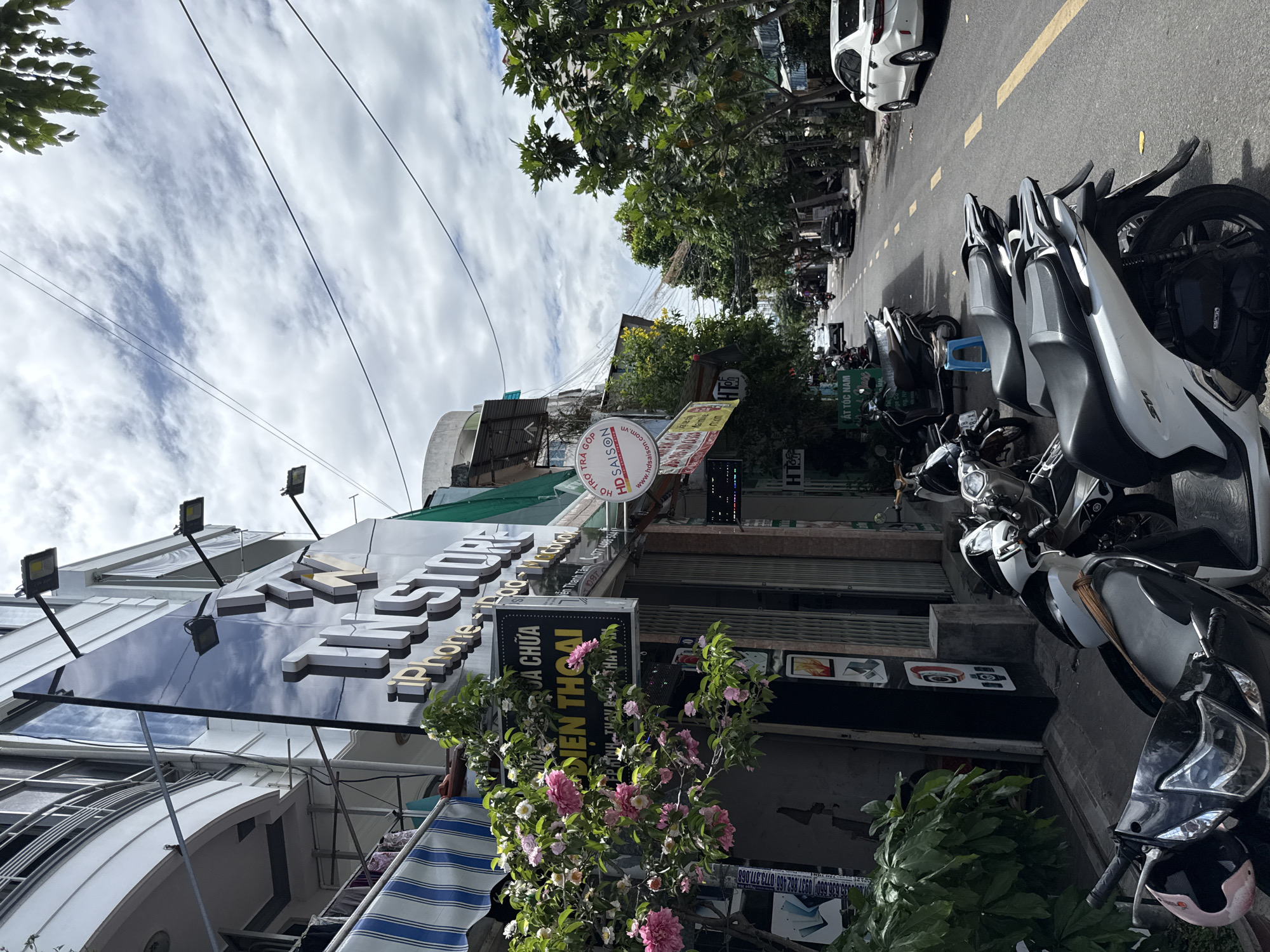 The image depicts a street scene in Nha Trang, Vietnam. On the left side of the street, a store called "TIN STORE" is visible, with signs indicating it sells iPhones and offers repair services for mobile phones. A flowering tree with pink and white blossoms is positioned in front of the store. Several motorbikes are parked along the sidewalk. The street itself is paved and lined with trees on the right side. Cars and other motorbikes are present on the street, indicating ongoing traffic. The sky is partly cloudy. The scene appears to be a typical urban commercial area.