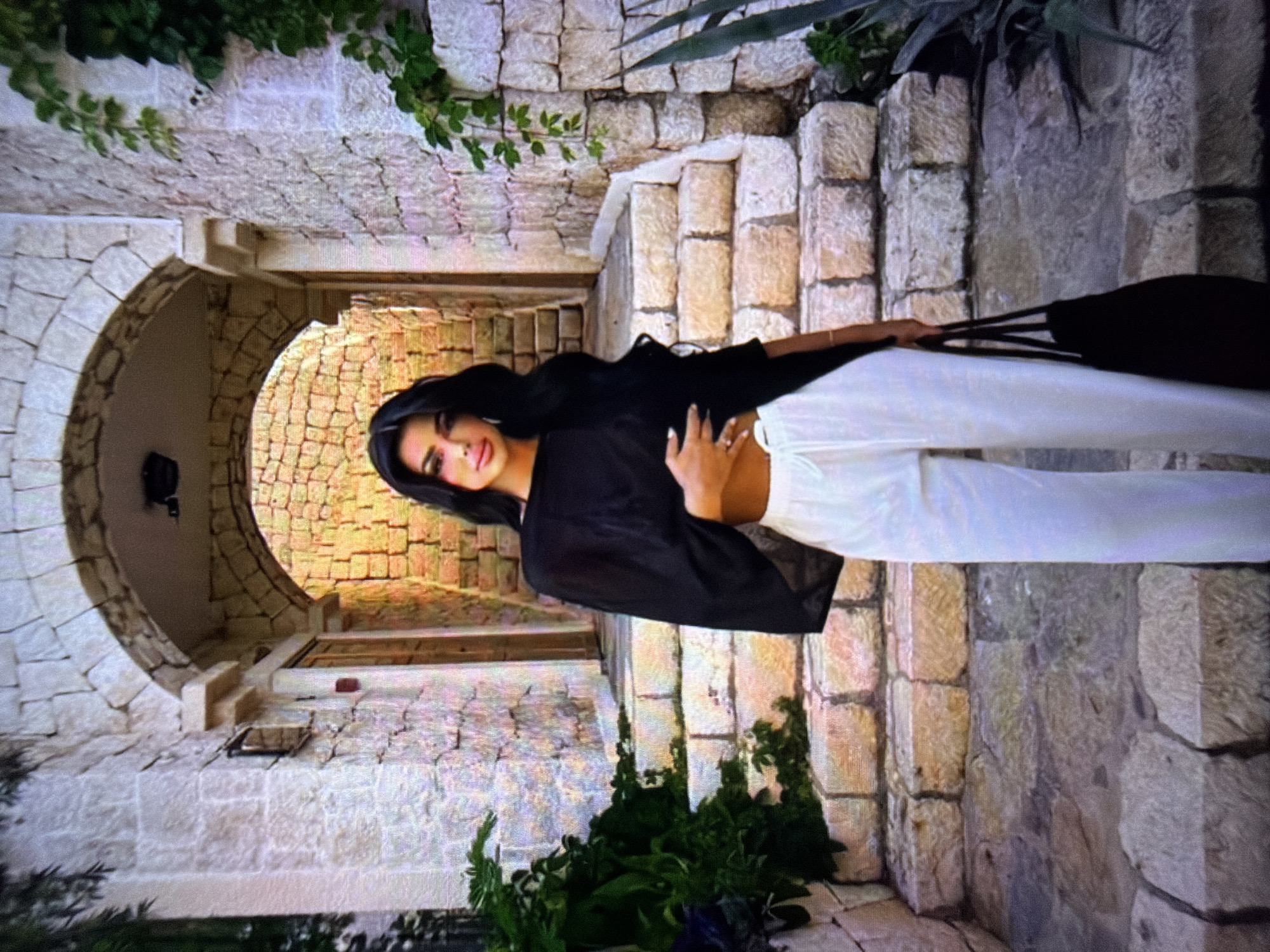 A woman stands on stone steps in front of an arched entryway. She has long dark hair and is wearing a black top and white pants. She is holding a black bag in her right hand. The entryway is made of stone, and there are plants on either side of the steps. The location is identified as Beșalma, Moldova.