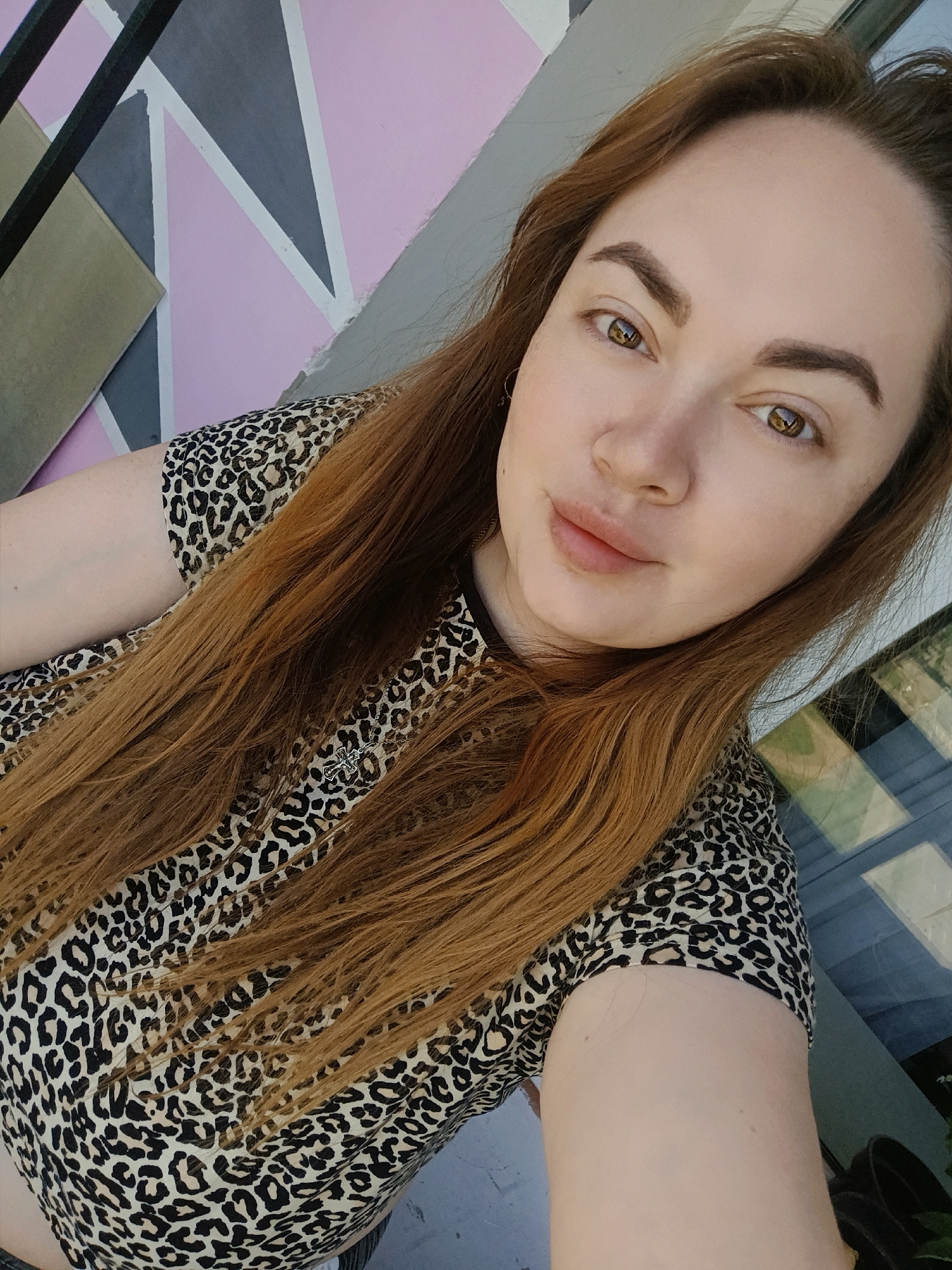 A medium close-up selfie of a woman with long, straight, reddish-brown hair, light skin, and defined eyebrows. She is wearing a leopard-print top and a silver necklace with a cross pendant. She has brown eyes. The background features a wall with geometric shapes in pastel pink, gray, and light beige, with a black metal railing partially visible at the top left. A window with reflective panes is to her right. The location is potentially Chaikovsky, Russia. No interactions or actions are visible.