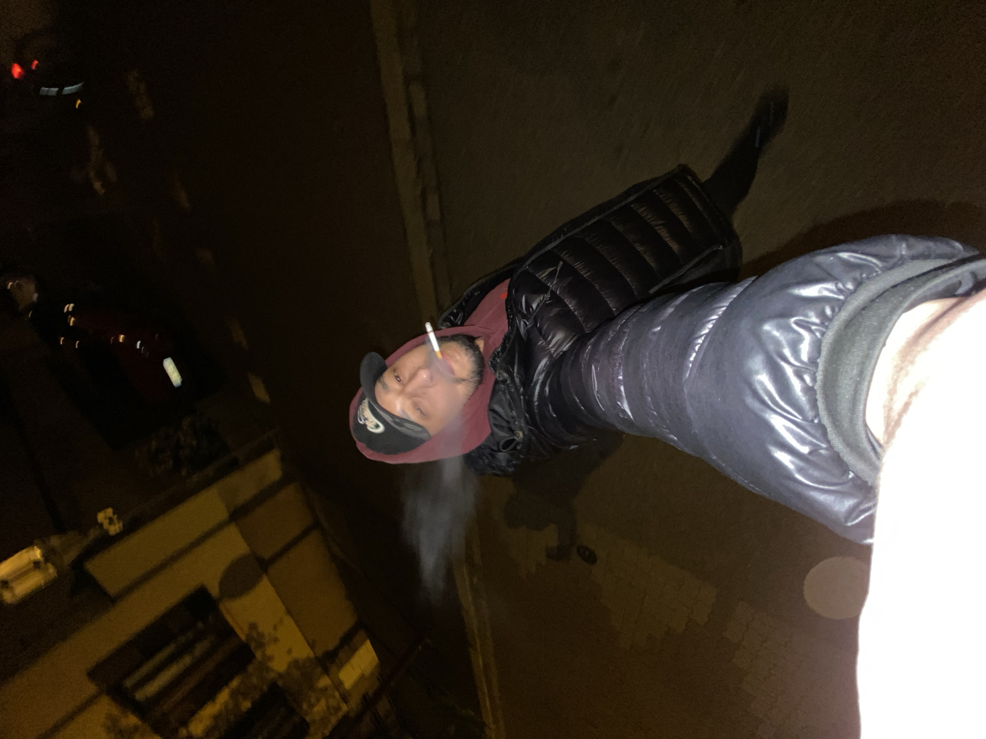 A person, angled from above, smokes a cigarette in an urban environment at night. They wear a dark, puffy jacket and a baseball cap. Smoke emanates from the cigarette. The setting appears to be a city street, with buildings and streetlights visible in the background. The location is in Schiltigheim, France.