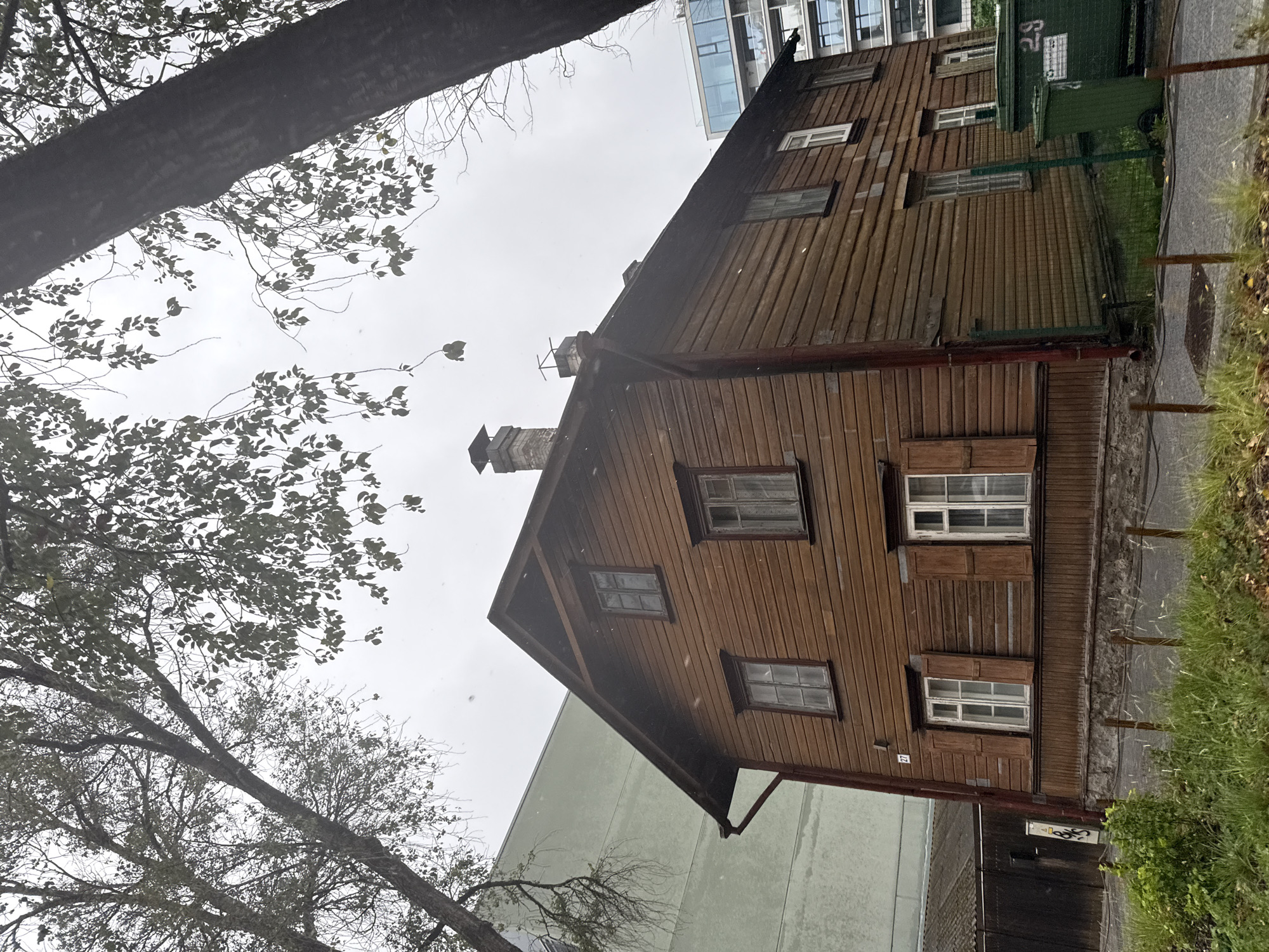A two-story wooden house with a dark brown facade and red trim stands in front of a modern building and a light green building, with large trees on either side. The wooden house has several windows on each floor. Some windows are open, revealing curtains. There are shutters on some of the lower windows. A brick chimney with two vent pipes protrudes from the roof. In front of the house, there is a chain-link fence and some green trash bins. The sky is overcast. The image appears to be taken on a street in Tallinn, Estonia.