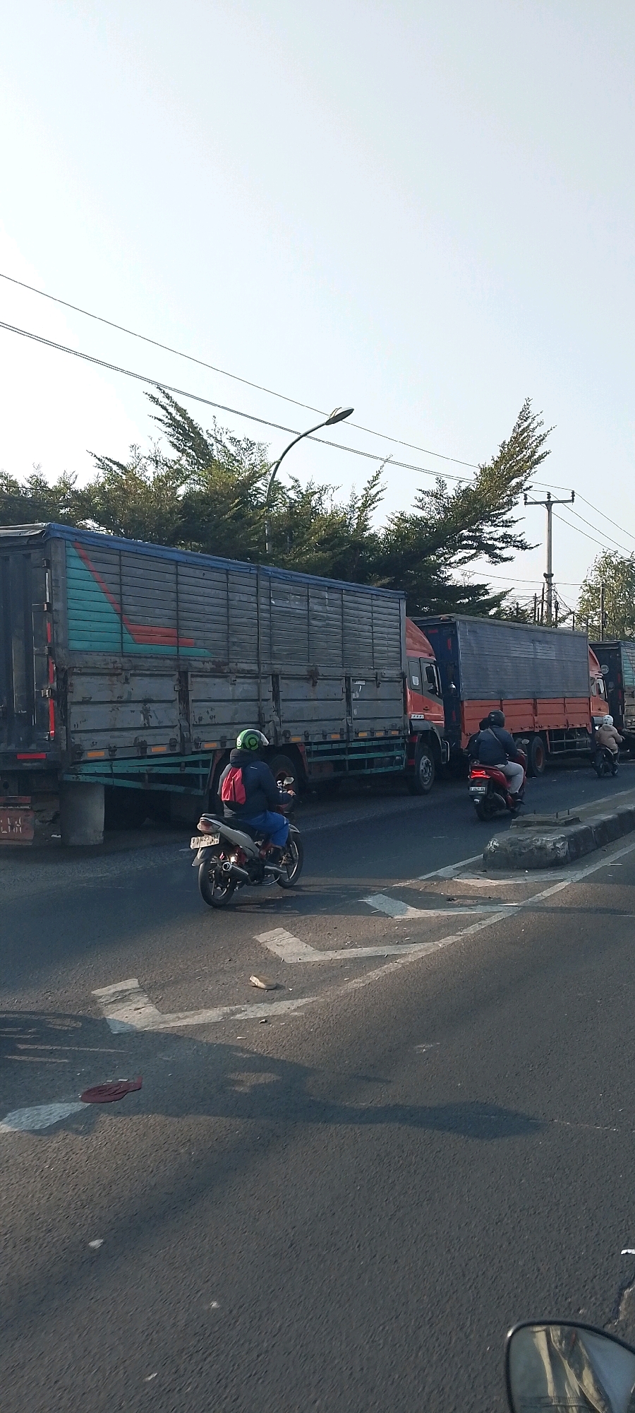 The image shows a street scene in Bekasi, Indonesia, with multiple large trucks and motorcycles. In the foreground, a person on a motorcycle wearing a green helmet and a red backpack is riding on the road. The motorcycle is silver with white and red accents. The road has white lane markings. Behind this motorcycle, there are several large trucks parked or moving slowly. The trucks are a mix of colors, including dark grey, orange, and blue. The sky is clear and blue, suggesting it is daytime. Trees and utility poles are visible in the background. The overall impression is of a traffic situation, possibly congestion.