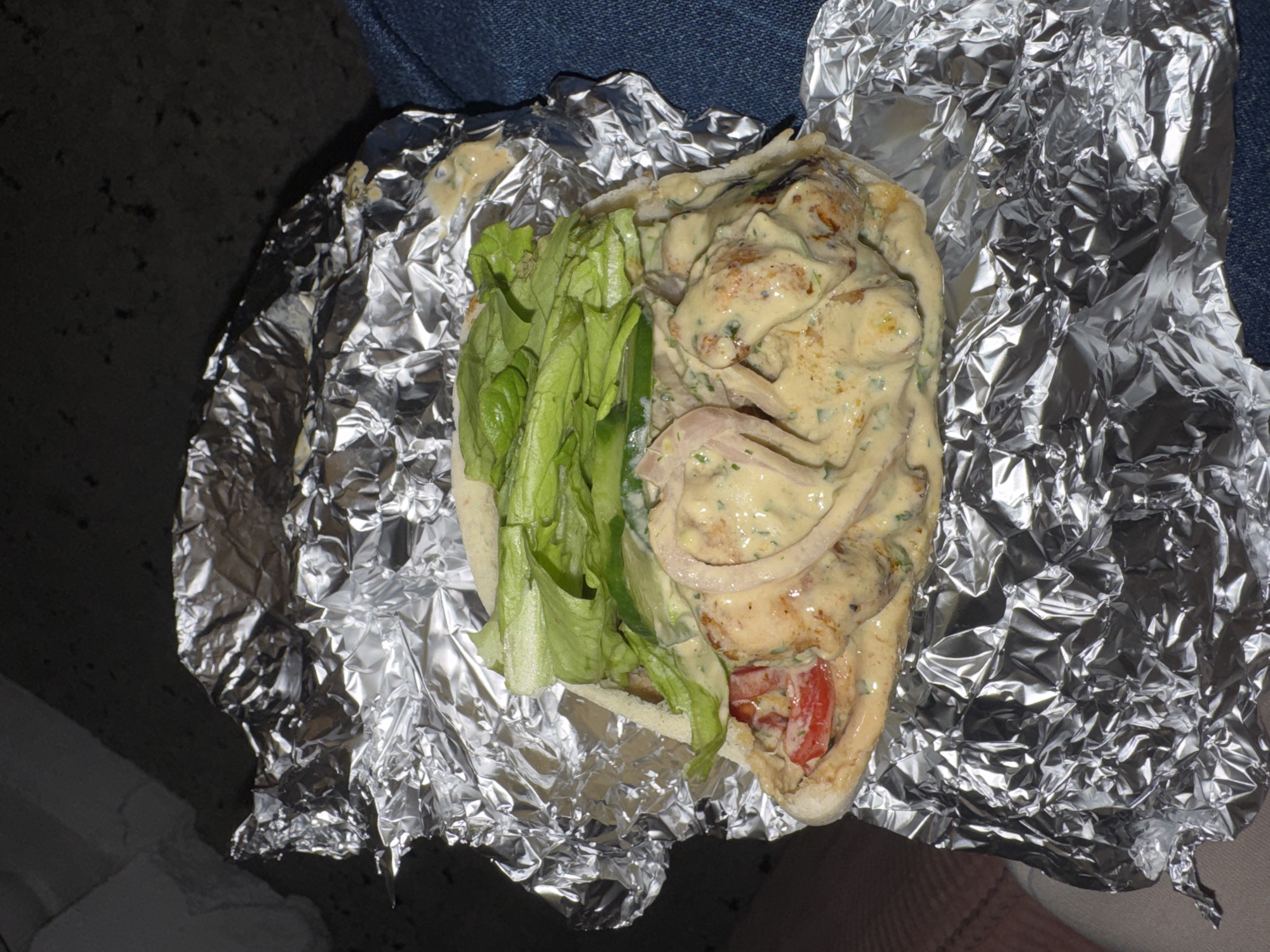 An overhead shot depicts a gyro sandwich unwrapped from aluminum foil. The sandwich contains sliced tomato, lettuce, chicken, and onion, all covered in a creamy sauce. The background consists of blue denim fabric and a textured gray surface. This image was taken in an unknown location, not necessarily in Tbilisi, Georgia.