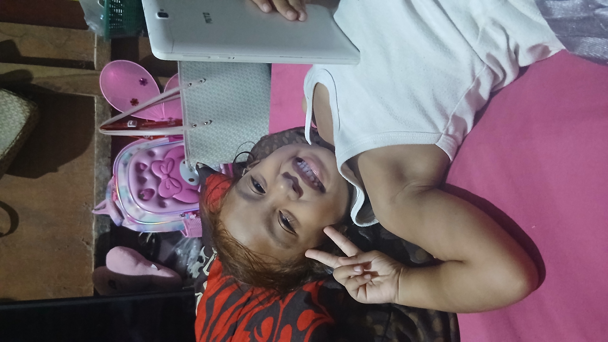 A young child, likely female, is lying on their back on a pink blanket, looking up at the camera. The child is wearing a white tank top. The child's head is tilted, and they are smiling with their mouth open, revealing their teeth. Their right hand is raised, with two fingers extended in a peace sign. The child's hair is dark and appears to be wet.
Behind the child, a white tablet is propped up, suggesting the child may have been watching something or playing a game. To the left of the child, a pink backpack with a cartoon character design is visible, along with a pink slipper. The background includes a wooden surface and a green basket. The overall setting appears to be indoors, possibly a bedroom or living area, given the presence of a blanket and toys. The lighting suggests it might be daytime, but it's difficult to ascertain the exact time. The child's expression conveys happiness and playfulness. The visible text on the tablet reads "MITO". The location context is Keudejungkagajah, Indonesia.
