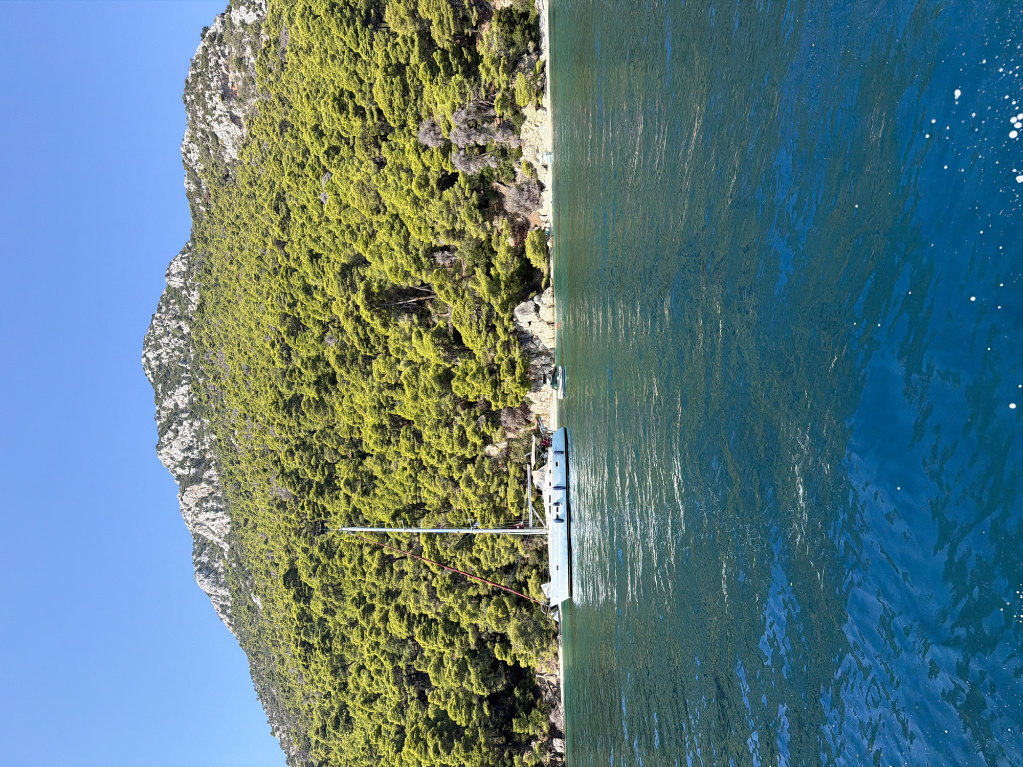 The image depicts a serene coastal scene in the Municipal Unit of Kalamos, Greece, under a clear blue sky. In the foreground, the deep blue, tranquil waters of the sea gently ripple. In the middle ground, a white sailboat with a red accent on its sail is anchored near a verdant, densely forested mountainside. The mountain is covered with lush green pine trees, with rocky outcrops visible towards its summit. Along the shoreline, a small beach with rocky sections is visible, with at least two other smaller boats moored near the shore. A few people can be observed on the beach. The overall impression is one of natural beauty and a peaceful vacation setting.