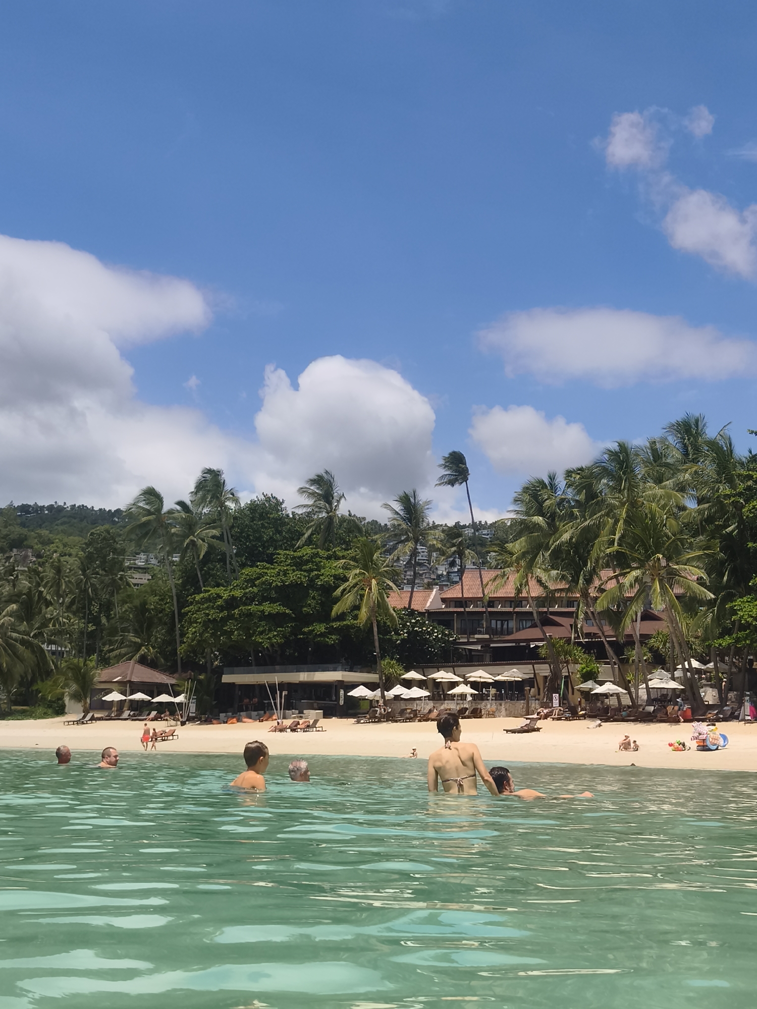 In the foreground of the image, clear turquoise water laps at a sandy beach under a bright blue sky with scattered white clouds. Several individuals are visible swimming in the water, some with their bodies partially submerged. In the middle ground, the sandy beach is lined with palm trees and lounge chairs with umbrellas, suggesting a resort or hotel setting. A building with a terracotta-tiled roof and multiple levels is visible behind the palm trees. People are seen relaxing on the beach. The background features a lush, green hillside covered in trees, indicating a tropical environment. The scene appears to be a vibrant and serene beach destination in Ko Samui District, Thailand, on a sunny day.