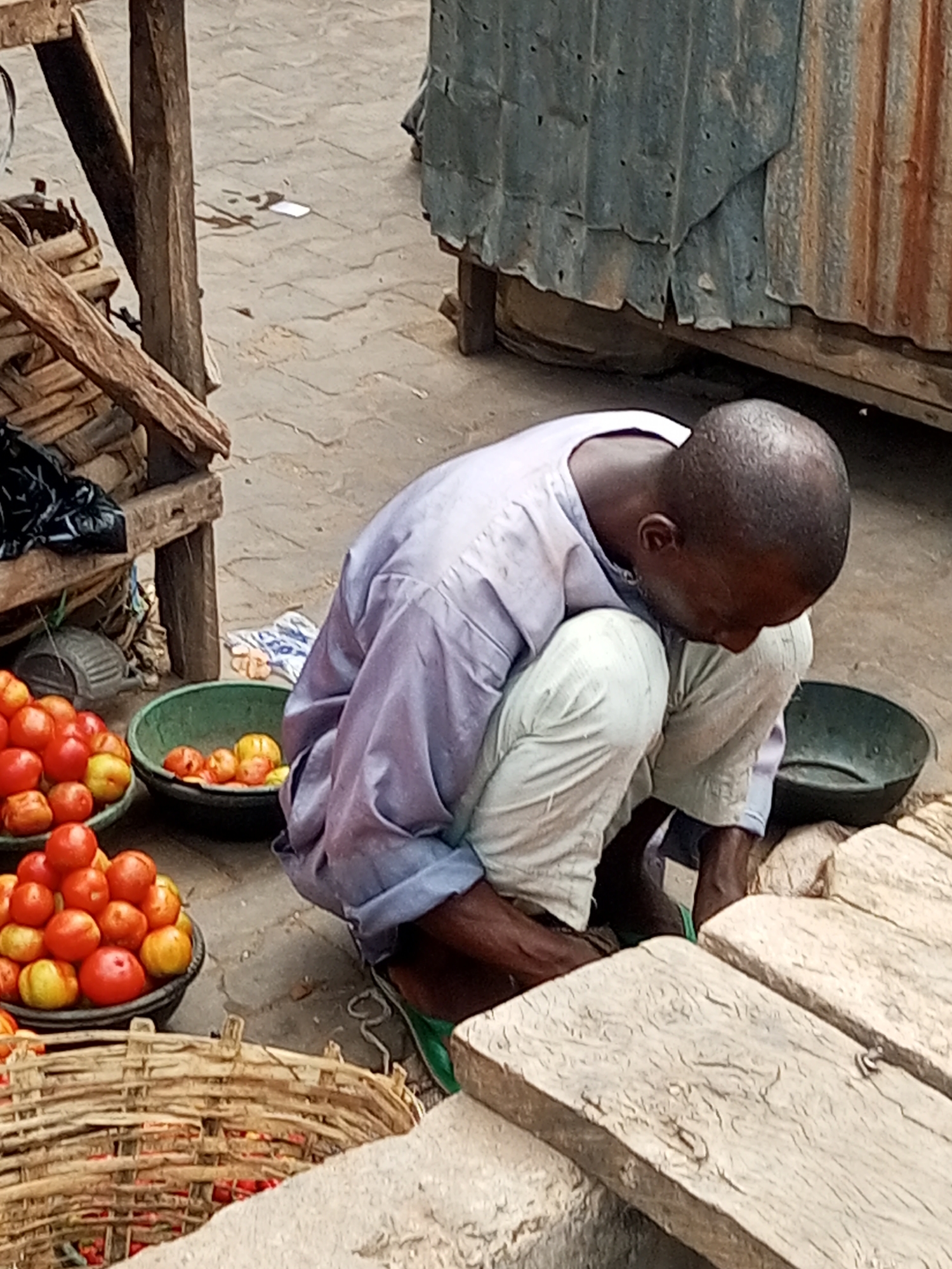 A person squats near a market stall in Tempure, Nigeria. The person wears a light-purple shirt and light-colored pants. Baskets and bowls filled with red and yellow tomatoes are visible on the ground. The market stall is constructed from wood and corrugated metal.