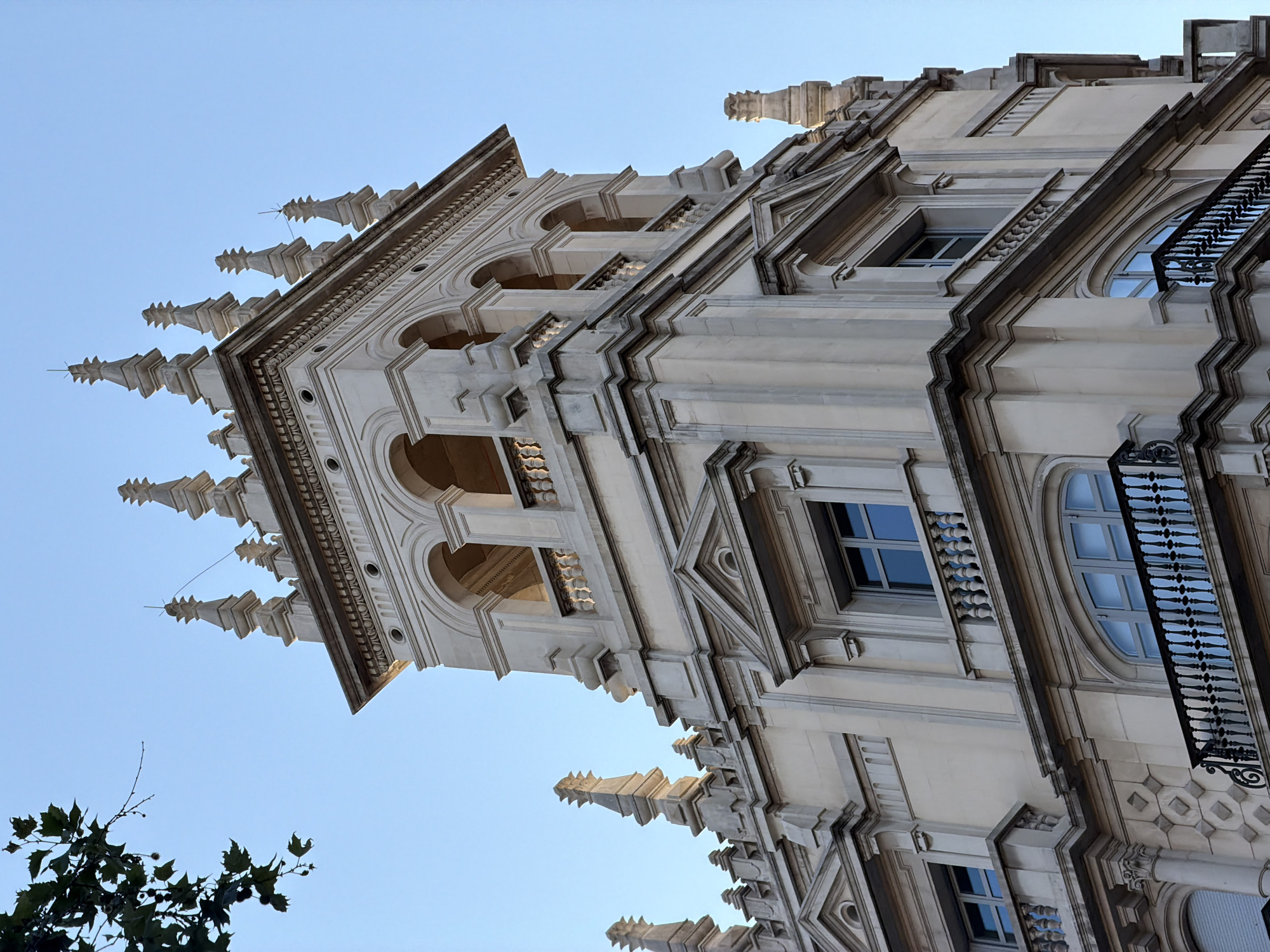 The image captures a close-up, low-angle view of a decorative stone building facade against a clear blue sky. The building features intricate architectural details, including arched windows, balconies with ornate balustrades, and a multi-tiered tower topped with spires. The stone appears weathered, suggesting an older structure. In the upper left corner, a branch with dark green leaves and some small seed pods hangs into the frame, providing a natural element against the man-made architecture. This building is located at Avinguda de Francesc Cambó, 2-12, 08003 Barcelona, Spain.