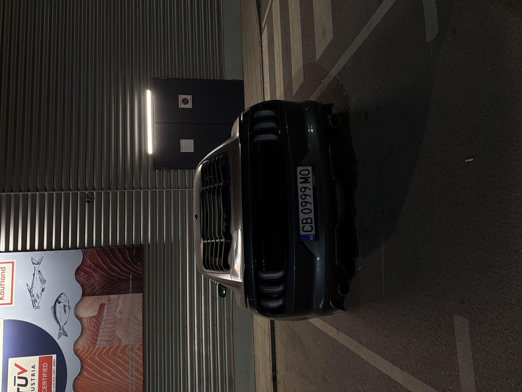 The image captures the rear view of a dark-colored car parked at night in what appears to be a commercial parking lot. The car is positioned on an asphalt surface, with white parking lines visible. To the left of the car is a vertical corrugated metal wall belonging to a building.
A bright, vertical fluorescent light fixture illuminates a section of the wall, highlighting two dark electrical panels mounted there. One panel clearly displays a "no entry" or "prohibited" symbol (red circle with a diagonal line).
Further left on the wall, a large advertisement poster is visible. It features illustrations of fish and cut salmon, indicating food products. Prominent text on the poster includes "Kaufland," "TÜV AUSTRIA CERTIFIED," and Bulgarian text: "ISO 9001, ISO 22000 и НАССР, което гарантира високи стандарти за качество, при съхранение на хранителните продукти." (translated: "ISO 9001, ISO 22000 and HACCP, which guarantees high quality standards, for the storage of food products.").
The car's license plate, "BG CB 0999 MO," identifies the vehicle as being from Bulgaria, with "CB" corresponding to Sofia. The time of day is night, evidenced by the artificial lighting and darkness. No people or specific activities are visible. The scene suggests a supermarket or large retail store parking area in Sofia, Bulgaria.