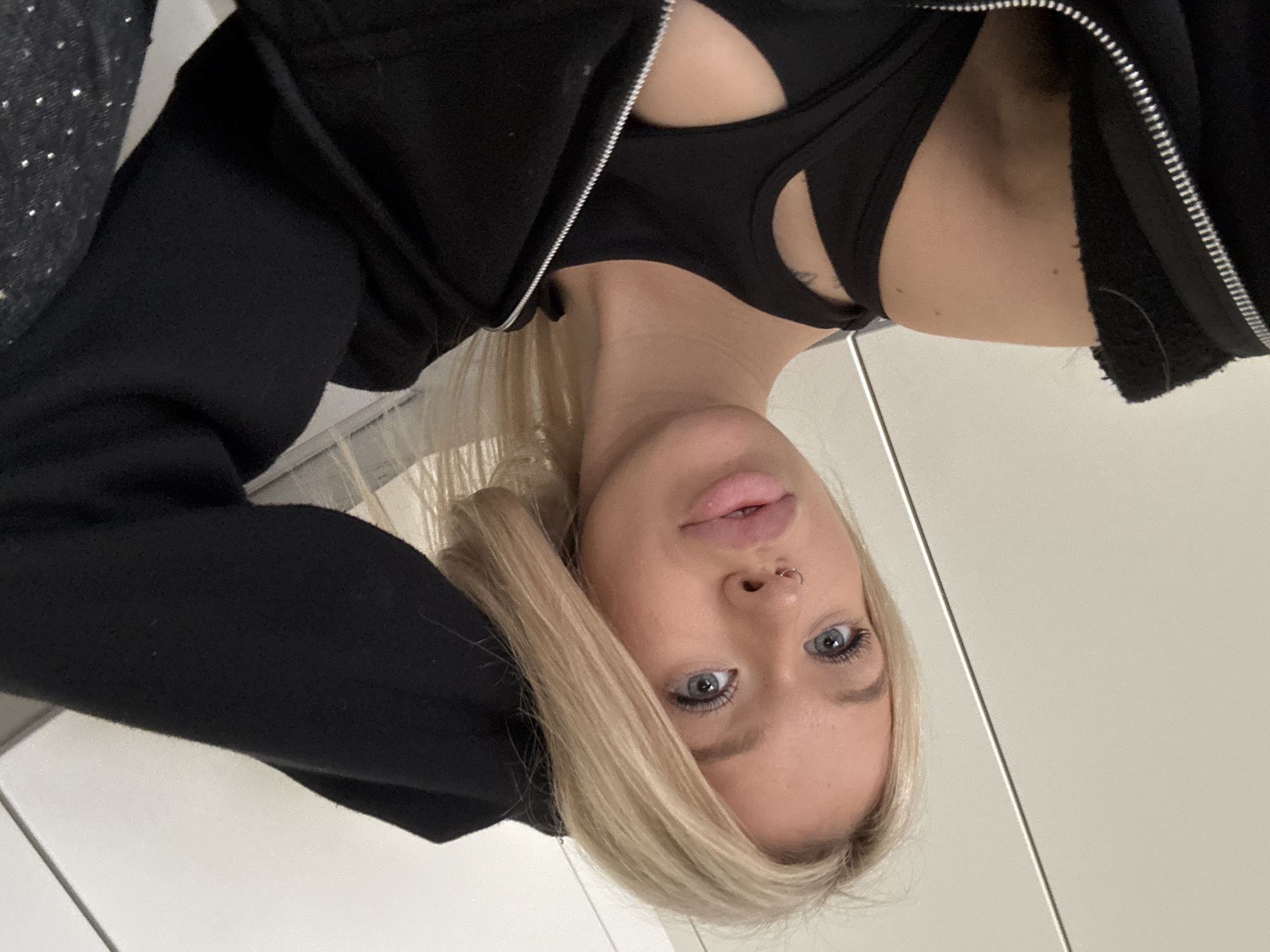 The media file is a portrait of a young Caucasian woman with blonde hair and blue eyes. She is wearing a black zip-up hoodie over a black sports bra. She has a nose ring and light makeup. The image is taken from a low angle, with the woman's face tilted towards the camera. Her expression is neutral.
The background appears to be indoors, possibly a room with light-colored walls. There is a hint of another surface or object to the left of the woman, which is dark and possibly textured with small reflective elements, suggesting it could be a garment or accessory. The lighting is soft and even, suggesting an indoor setting with artificial light. There are no clear indications of time of day or weather conditions. The location context of Saint Petersburg, Russia, does not offer any readily discernible visual cues within the image itself. There is no visible text in the media file. The activity appears to be a casual selfie or posed portrait.