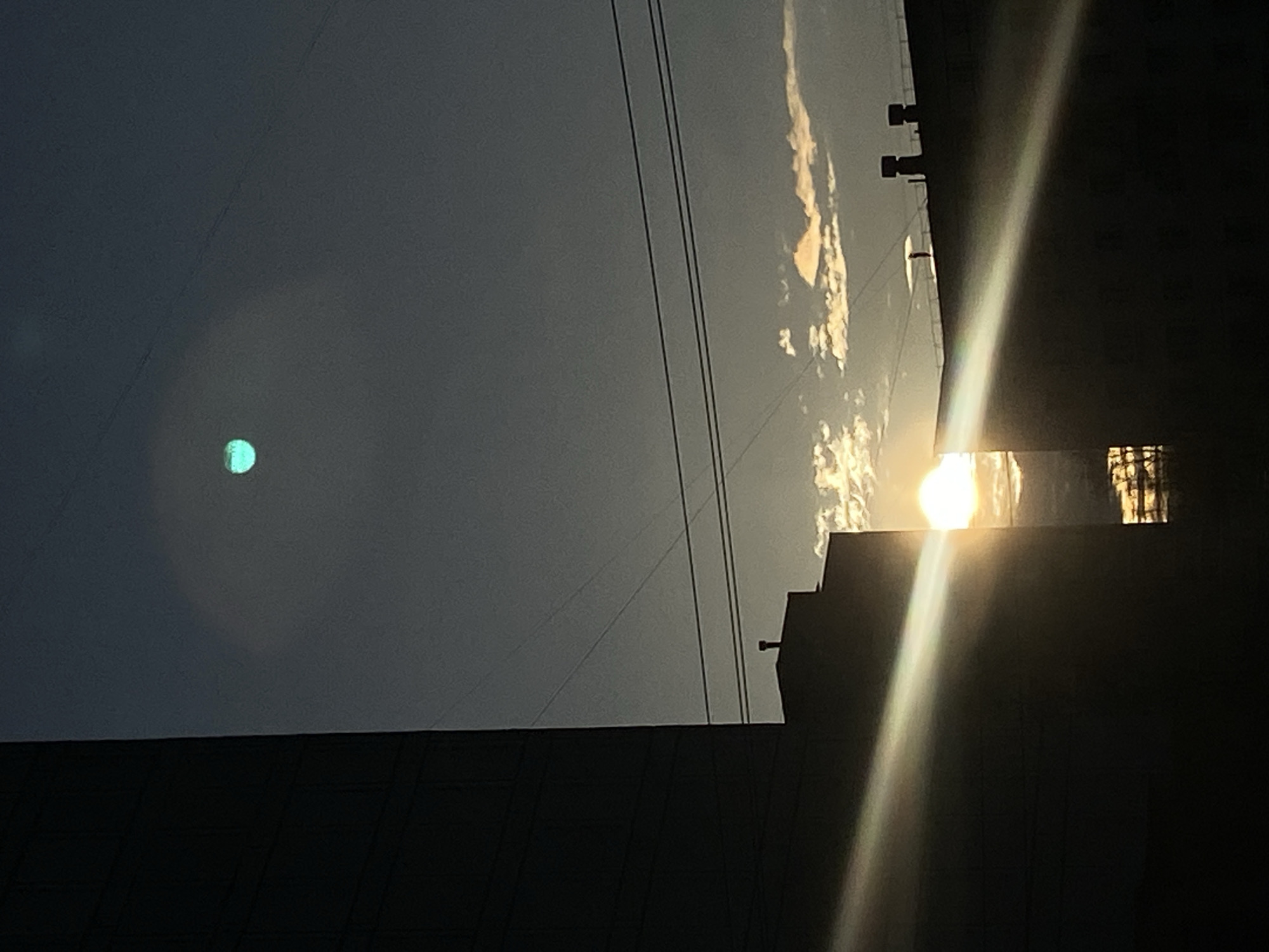 The image shows a view in Chaikovsky, Russia. The sky is gray, with what appear to be utility wires crossing the upper part of the frame. The sun is visible, creating a bright glare with a white beam. The sun is partially blocked by a dark building. A small, turquoise-colored object is visible in the sky to the left of the sun. The bottom portion of the frame is occupied by dark buildings.