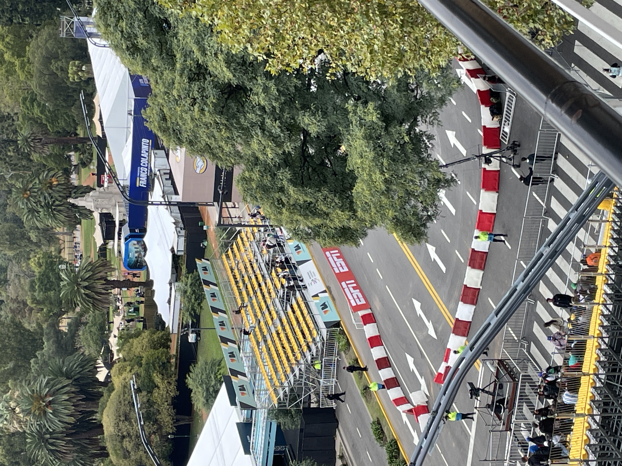 This image shows a section of what appears to be a temporary street circuit in Buenos Aires, Argentina. The circuit has barriers of red and white, with yellow arrows indicating direction of travel on the track. Spectator stands line one side of the track and are filled with spectators, whilst others are visible along the barricades. A green area with various trees and plants borders the other side of the track. There are many people in attendance. The image suggests a racing event.