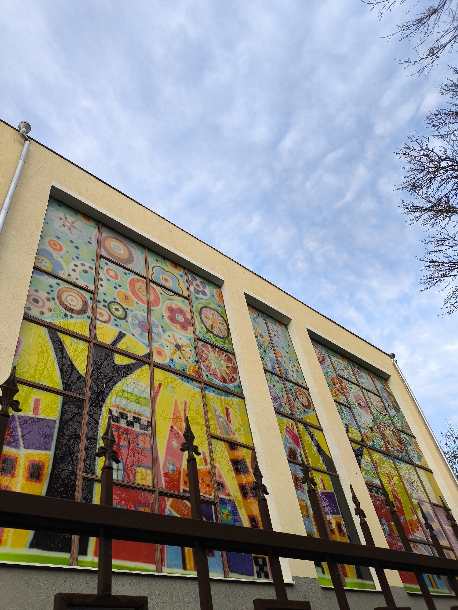 A low-angle, medium shot captures the exterior of a building with a large, brightly colored stained-glass window, viewed from behind a dark metal fence. The building facade is a pale yellow, with the stained-glass window dominating the center of the frame. The window is composed of numerous rectangular panes, each filled with vibrant, abstract patterns featuring circles, flowers, and geometric shapes in a variety of colors, including yellow, blue, red, purple, and green. Reflections of trees and the sky are visible within the lower panes of the stained-glass, suggesting it is daytime and that the window faces an outdoor area. The sky above is partly cloudy with a light blue hue, and the bare branches of a tree are visible in the upper right corner. The fence in the foreground consists of dark, ornate metal bars, some with pointed finials, suggesting a boundary to the property. The overall scene conveys an artistic and somewhat whimsical aesthetic, with the stained-glass window serving as a focal point. This image is of the stained-glass window of the Yessentuki Railway Station in Yessentuki, Russia.