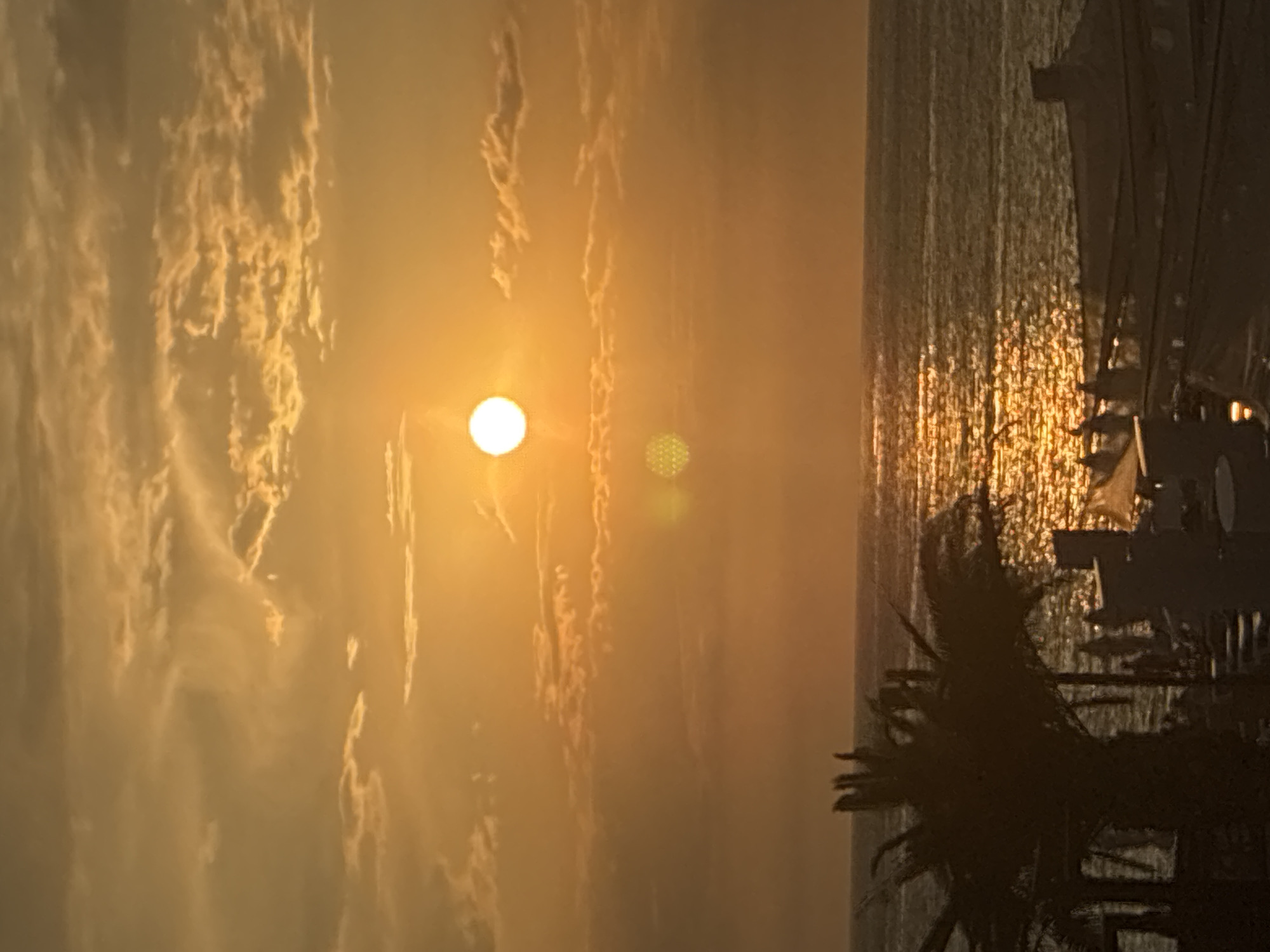 An aerial view captures a sunset over the Black Sea in Sochi, Russia. The sun, a bright white orb, is low in the sky, casting a warm, golden glow on the scattered clouds and the rippling water. The reflection of the sun creates a shimmering path across the sea's surface. In the foreground, silhouetted against the vibrant sky, are palm trees and structures that suggest a resort or beach area, with several indistinct figures visible amongst them. The scene evokes a peaceful, end-of-day atmosphere.