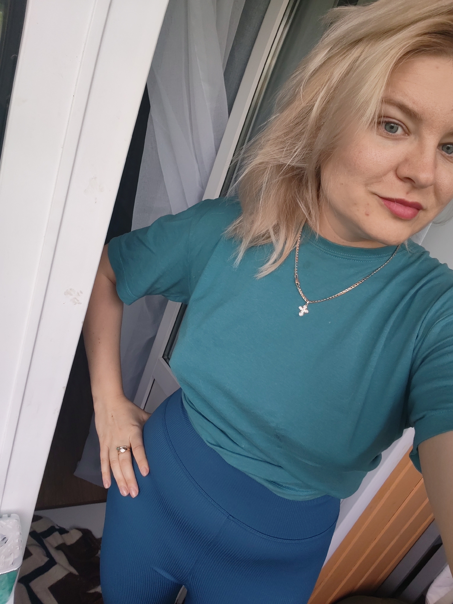 A Caucasian woman with blonde hair, blue eyes, and light skin poses for a selfie. She is wearing a teal t-shirt and blue leggings, with a gold cross necklace. A ring is visible on her left hand, which is placed on her hip. The background suggests an indoor setting with a white door frame and sheer white curtains. The woman appears to be in a residential area or apartment. There is no indication that the scene is located in the city of Izhevsk, Russia.