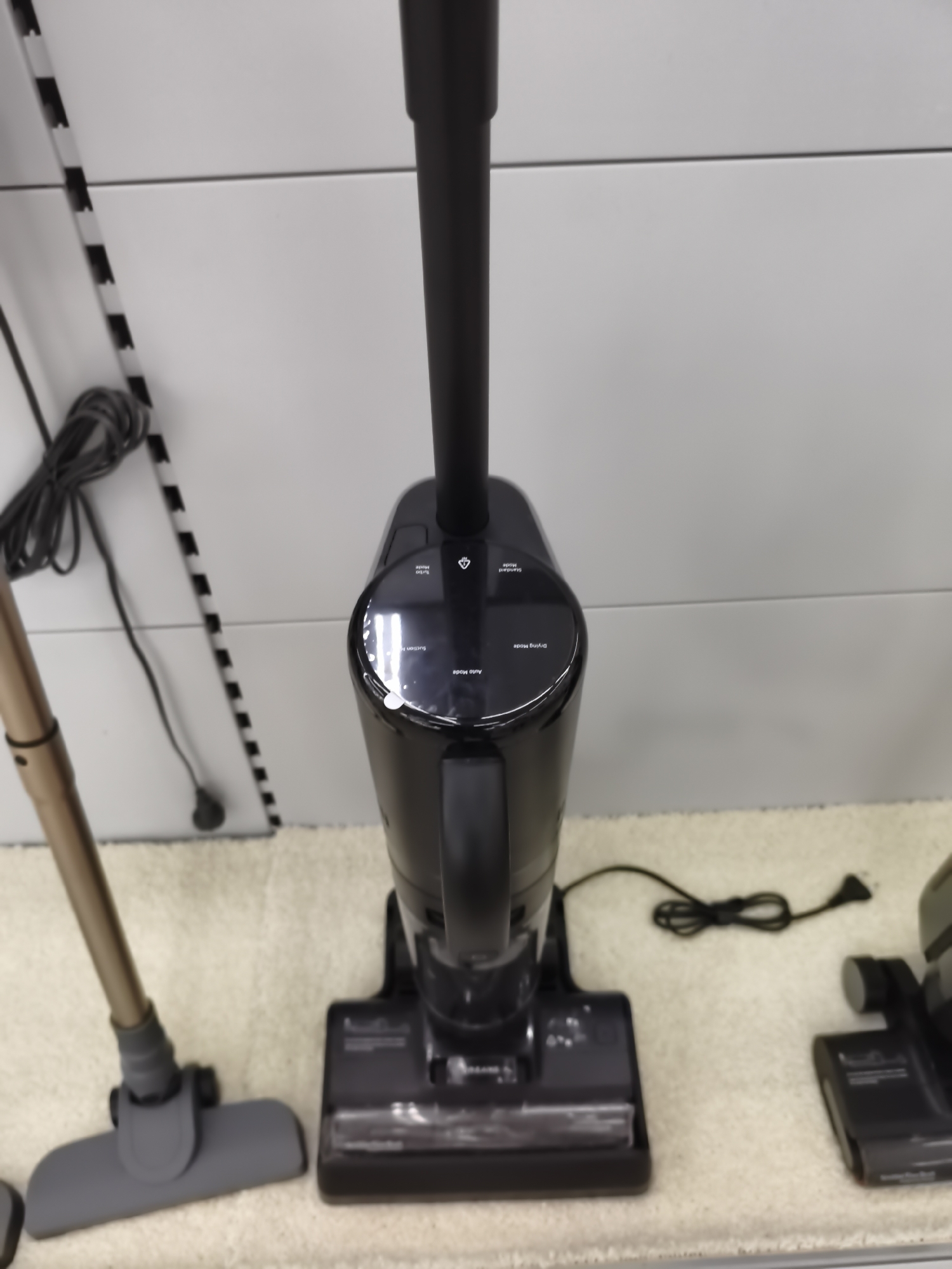 The image shows a close-up, eye-level view of a black stick vacuum cleaner placed on a beige carpet. The vacuum is upright, with its handle extending upwards out of the frame. The base of the vacuum is visible, featuring a cleaning head and a control panel with illuminated icons and text indicating different modes such as "Drying Mode," "Auto Mode," "Suction Mode," and "Standard Mode." To the left of the main vacuum, the brush head and lower portion of a second vacuum cleaner, with a golden-brown pole, are visible. The background consists of a light-colored, textured wall with horizontal lines and a striped black and white object leaning against it on the left. A black power cord is visible on the carpet behind the main vacuum, plugged into an unseen outlet. The scene appears to be an indoor retail display or storage area. The city of Aktobe, Kazakhstan is not depicted in this image.