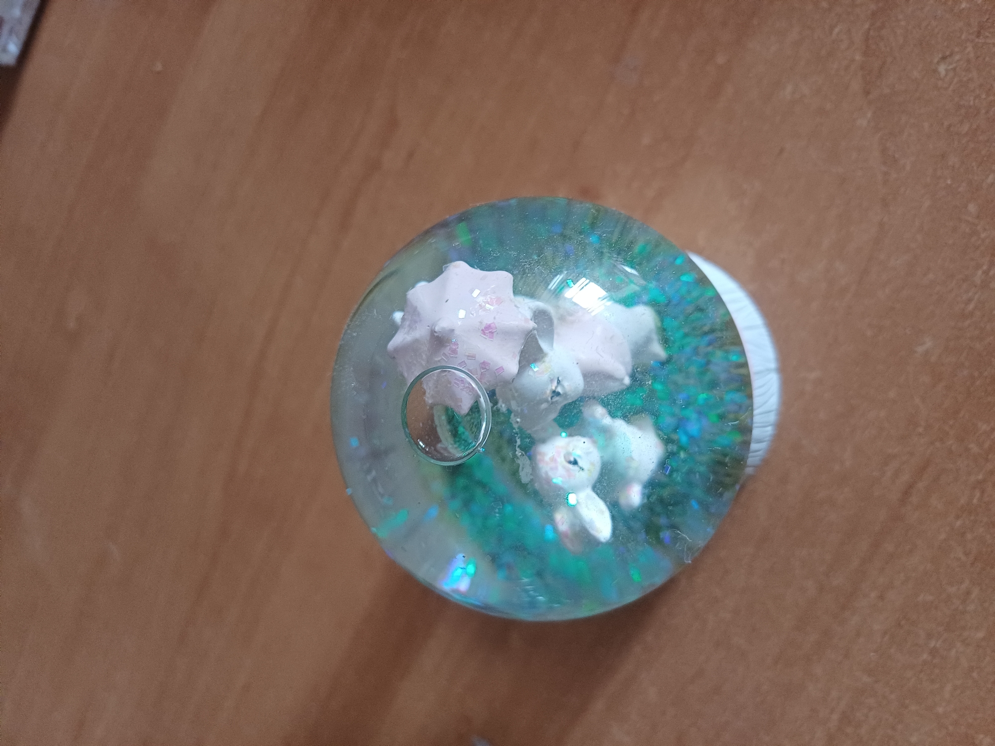 A snow globe sits on a wooden surface. Inside the snow globe are two small white figures, possibly rabbits, under a pink umbrella. The globe is filled with a blue liquid and glitter. The wooden surface appears to be a table. The background is a blurred brown surface. The scene is indoors, and no specific location in Kyiv, Ukraine is visible.