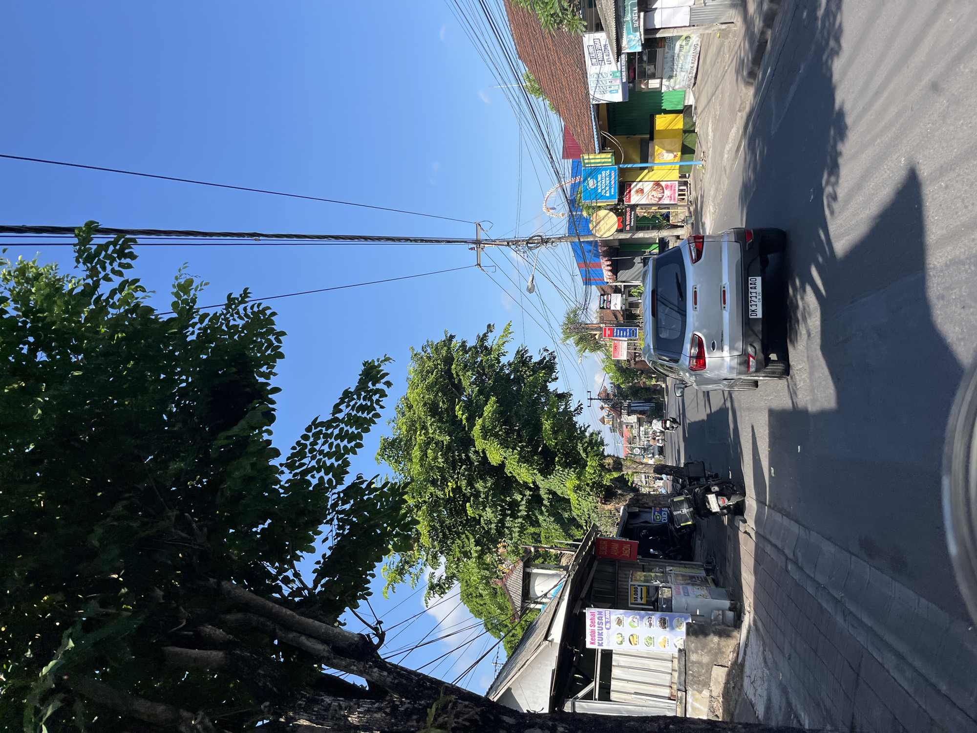The media file captures a street scene in North Kuta, Indonesia, under a bright, clear blue sky, suggesting a sunny day, likely mid-morning or afternoon. The perspective is from a vehicle, looking towards a roadside with shops and trees.
The foreground is dominated by the left side of a car, with its license plate clearly visible as "DK 1711 AAO". A large, leafy tree with a thick trunk occupies the left side of the frame, casting shadows. Power lines crisscross the sky, typical of many Southeast Asian streetscapes.
The roadside is lined with small shops and stalls. Some feature colorful signage, with text visible in Indonesian. One sign appears to advertise "Kedai Sehat KUKUSAN" with prices like "Rp. 2.000" and "10.000", indicating a food or drink establishment. Another sign mentions "LOW CALORIE", "ADDED SUGAR", and "4.8% ALCOHOL", possibly referring to beverages. Businesses like "RUNG BUK KOMANG" are also indicated. Motorcycles are parked along the sidewalk, and a few are visible on the road.
A silver SUV is the most prominent vehicle, driving down the street. The overall impression is a bustling local street with typical daily activities, commerce, and infrastructure. The bright sunlight and clear sky create a vibrant, tropical atmosphere. There are no discernible people in clear focus, but the presence of vehicles and shops implies the presence of people in the scene.
