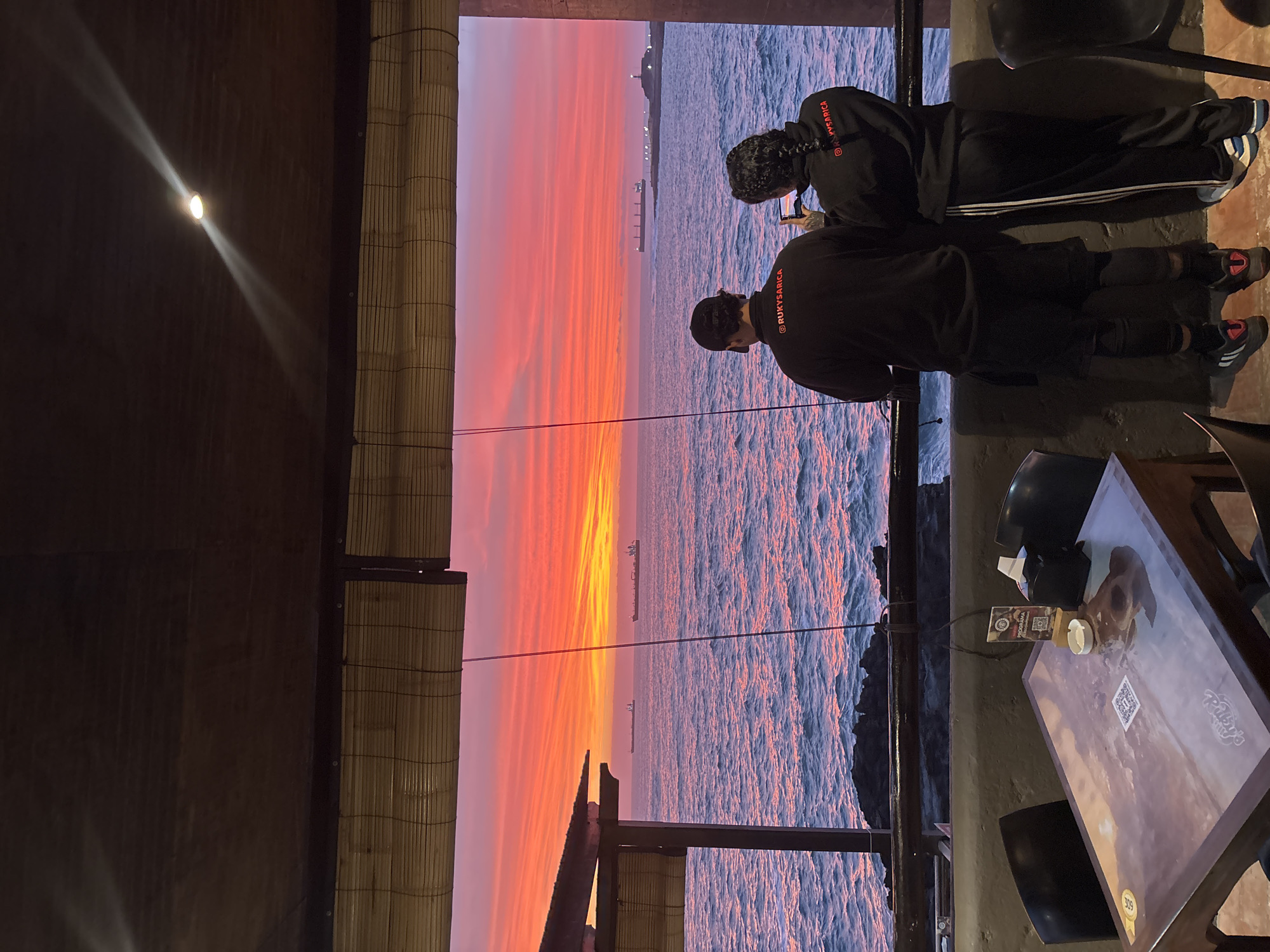 The photograph shows two individuals looking out at a sunset over the ocean from what appears to be a bar or restaurant setting. They are both wearing dark-colored clothing with "RUKYSARICA" written on them, and one of them is using a mobile phone, possibly taking a picture of the view. The horizon is visible through the structure of the bar, including a body of water with ships and a vivid orange and pink sunset. A table with items on it, including packaging from the restaurant and chairs are also in the frame. The photograph has been taken from an elevated angle, looking down towards the view. The scene is likely in the city of Arica, Chile given the writing on the clothing.