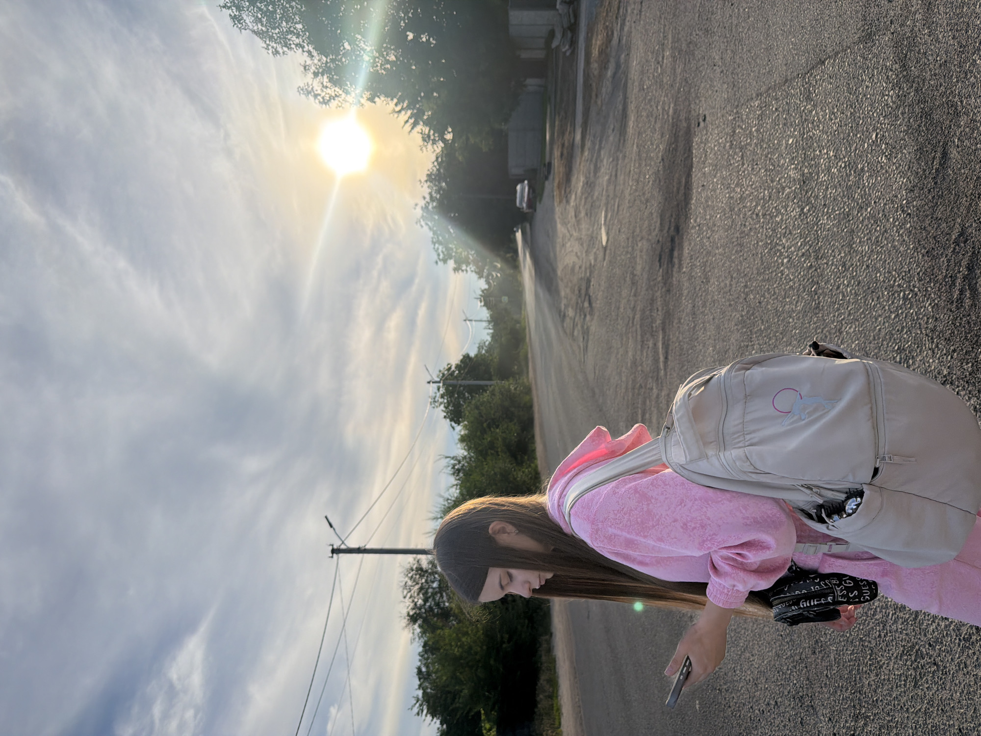 A young person with long brown hair, wearing a pink tracksuit and a beige backpack, stands on an asphalt road. They are holding a smartphone in their left hand and looking down at it. The sun is shining brightly in the sky, casting long shadows. Trees and power lines line the road, and a few houses are visible in the background. The scene appears to be a quiet, suburban street.