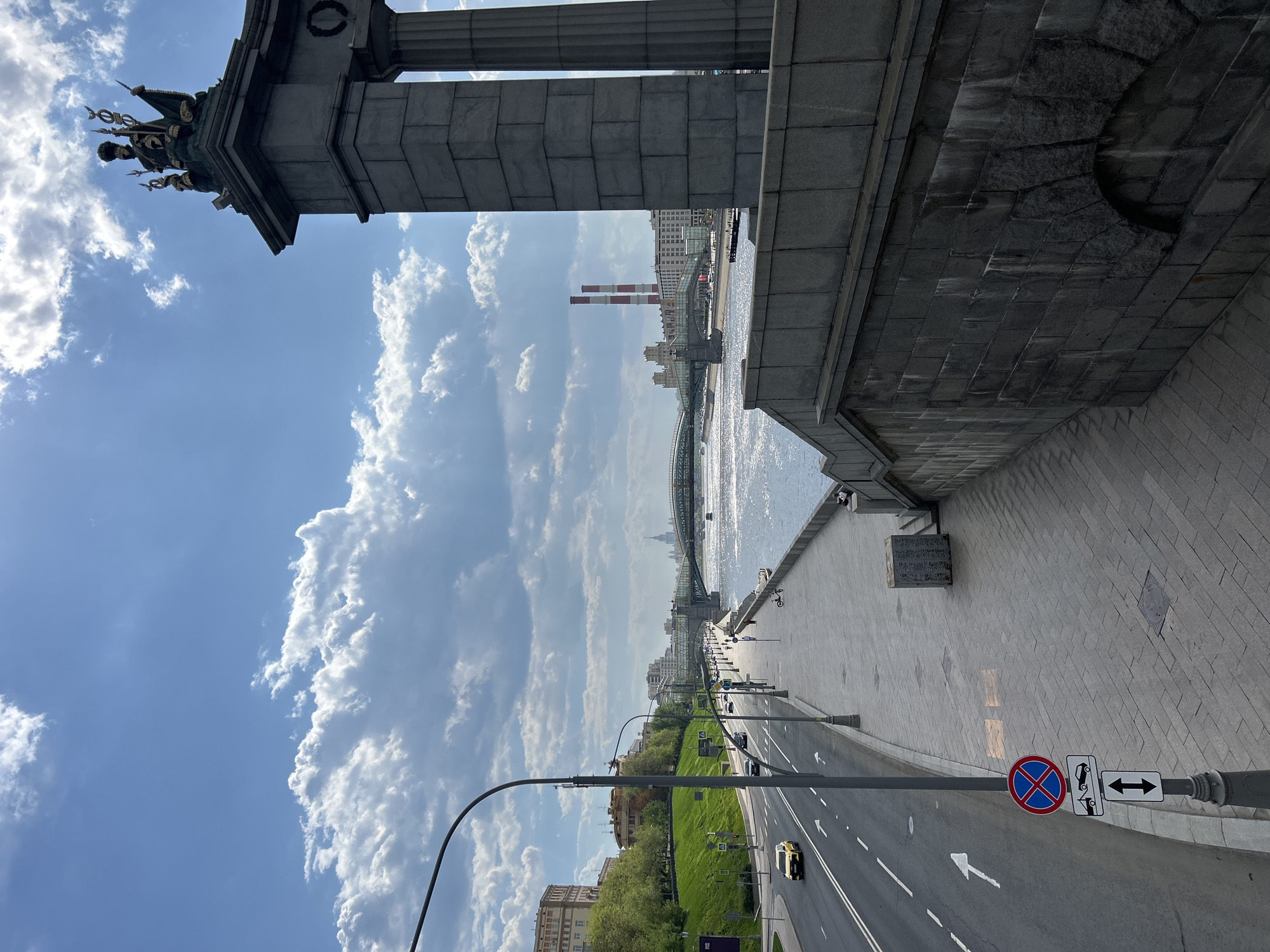 The image depicts a street scene in Moscow, Russia, under a bright blue sky with scattered clouds. The perspective is from an elevated position, possibly a bridge or high walkway. The view shows a road with white lane markings and a yellow car driving away from the viewer. There is a stretch of green space on the side of the road. The background features a body of water with a bridge spanning it. Beyond the water, city buildings and two tall red-and-white smokestacks are visible. In the foreground, part of a stone structure is prominent, including columns and a sculptural element at the top. A road sign indicating "no stopping" and a directional arrow are visible on a pole near the bottom right. The scene is urban, with elements of infrastructure, nature, and architectural details.