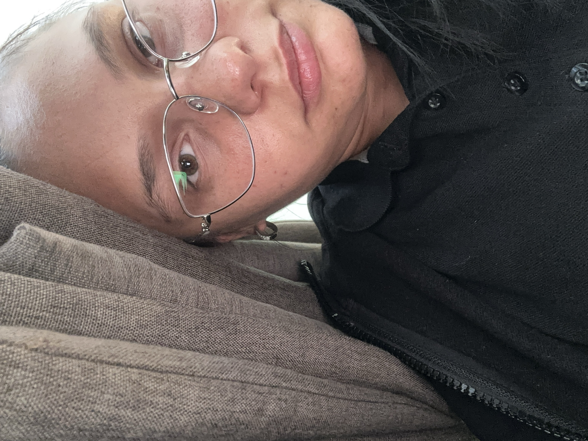 A close-up selfie shows a woman with light skin and brown eyes, wearing silver wire-frame glasses. She is lying on a grey-brown fabric surface, possibly a couch or bed. She is wearing a black zip-up jacket or sweater. The image does not provide information about the location, Sofia, Bulgaria, or any actions or interactions.
