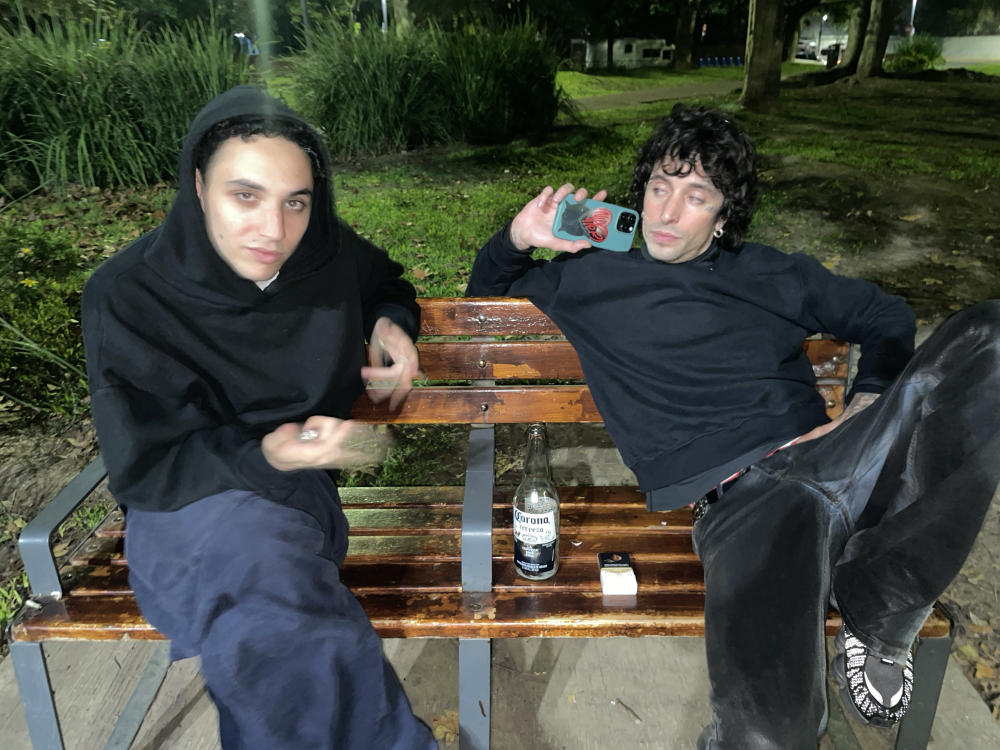 Two young men are sitting on a wooden bench in a dimly lit outdoor area. The man on the left is wearing a black hooded sweatshirt and navy blue pants. The man on the right is wearing a black long-sleeved shirt and black pants, and has a smartphone in his left hand. On the bench between them are a bottle of Corona beer and a pack of cigarettes. The background shows trees and vegetation, suggesting a park. The location is likely Buenos Aires, Argentina, based on the style of the street lamp and the clothing styles.