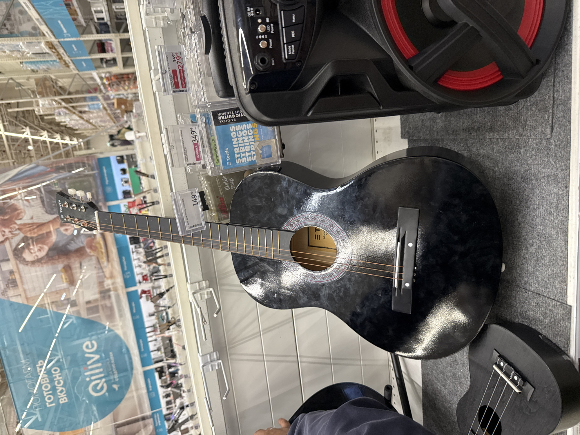 The image shows a retail store in Khimki, Russia, likely a consumer electronics or musical instrument store. In the foreground, a black acoustic guitar is prominently displayed, leaning against a white shelving unit. To the right of the guitar is a black portable speaker with red accents. In the lower left corner, a portion of a smaller, black stringed instrument, possibly a ukulele, is visible. Behind the guitar, shelves are stocked with various items, including what appear to be guitar strings and accessories. A large blue banner hangs in the background with Russian text and the logo "Qilive". The environment is well-lit and appears to be a typical retail setting with clean floors and organized displays. No people are clearly visible in the frame.