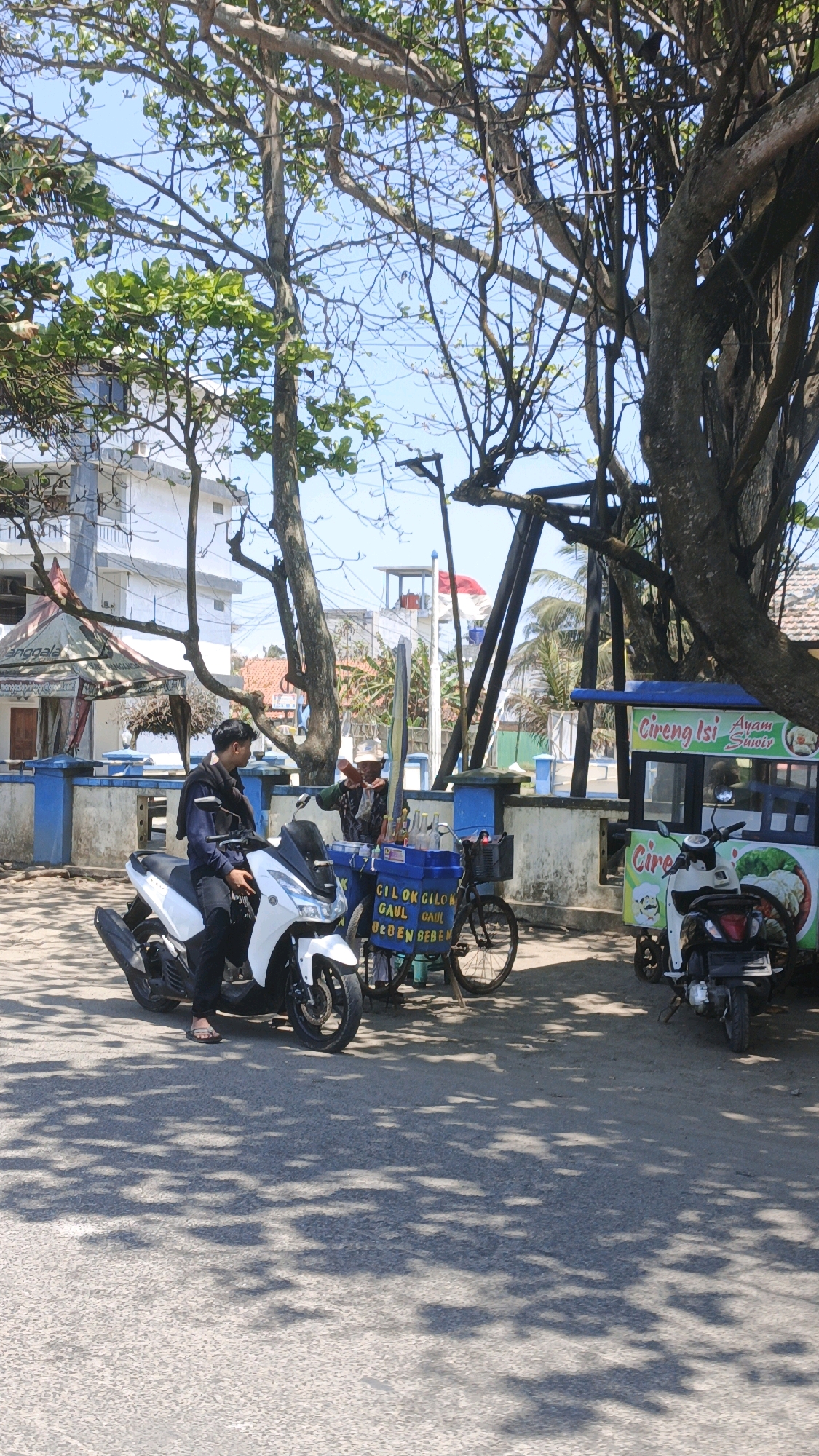 A man in dark clothing and flip-flops sits on a white scooter parked on a paved surface in front of a blue-and-white wall. He is looking towards a food cart where another man wearing a hat and a patterned shirt is serving a customer, who is out of frame. The food cart has bottles of drinks and what appears to be ice inside. The cart has text written on it, including "CILOK CILOK GAUL BEBEN BEBEN". Next to the cart is a bicycle with a basket. To the right of the cart is a smaller cart advertising "Cireng Isi Ayam Suwir". A white scooter is parked next to this cart. The scene is set under the dappled shade of large trees. In the background, there is a white building with blue accents and a white and red Indonesian flag flying. The sky is clear and blue. The location is identified as Desa Pagandaran, Indonesia, a coastal area, implied by the presence of a beach and vegetation typical of a tropical environment.