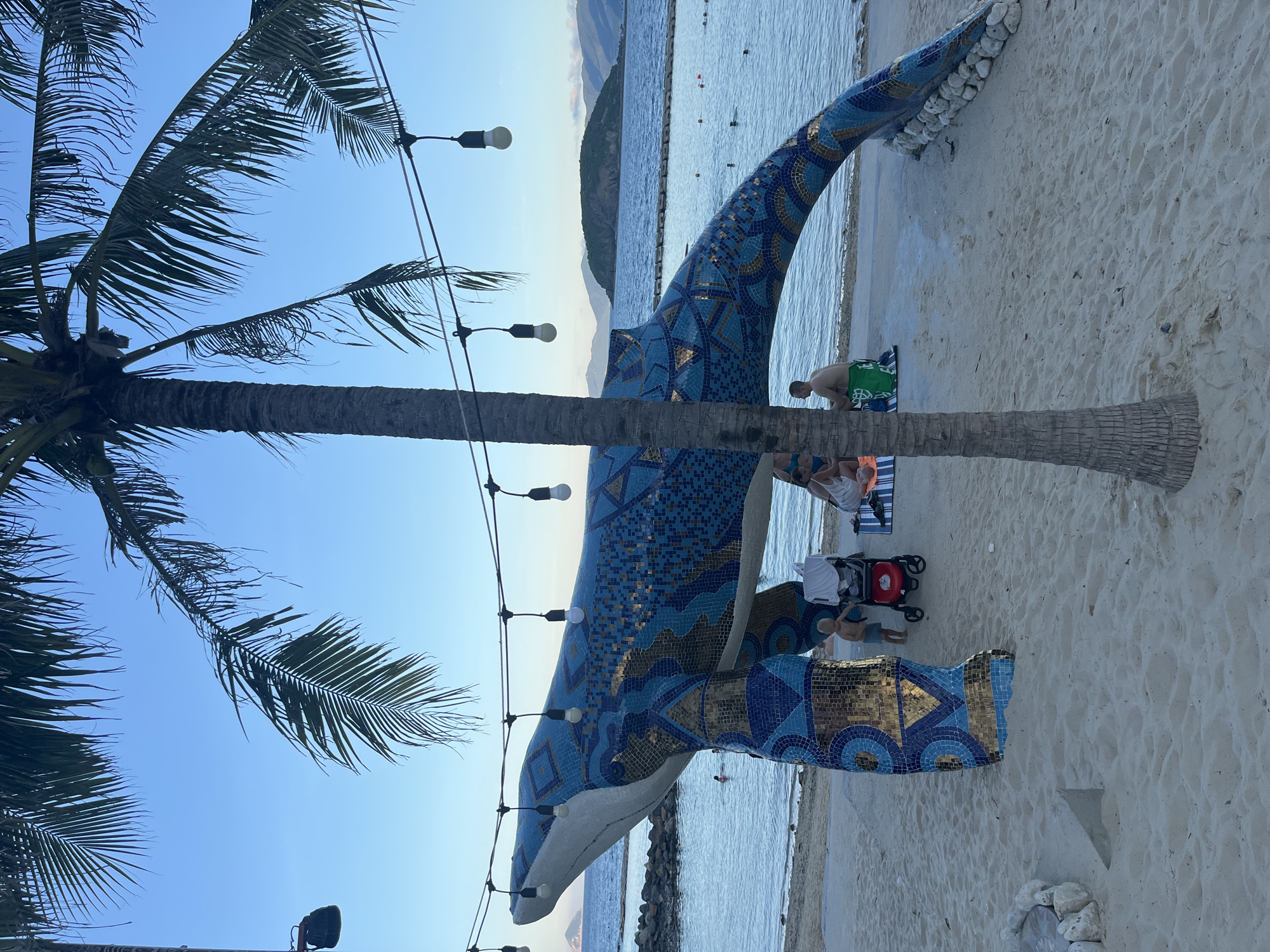 An outdoor, beachfront scene features a large mosaic sculpture of a whale. The sculpture is predominantly blue with intricate gold and darker blue patterns. It stands on a sandy beach, with its body arching over the sand, supported by its tail and head. A palm tree trunk is positioned in the foreground, partially obscuring the whale sculpture. String lights with exposed bulbs hang across the scene above the whale.
In the mid-ground, several people are visible. A young child stands next to a red stroller near the base of the whale sculpture. Further back, two adults are seated on a striped mat on the sand. Beyond the beach, the ocean stretches out, with several people swimming or wading in the shallow water. A rocky breakwater runs parallel to the coastline, and in the distance, a small, vegetated island and rolling hills are silhouetted against a clear blue sky. The overall ambiance suggests a relaxed, tropical resort setting during daylight hours.