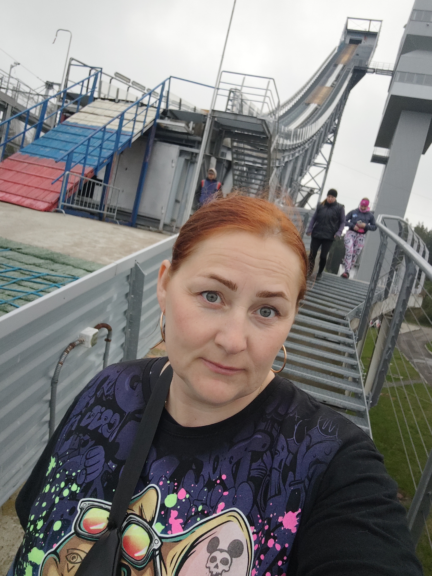 The image shows a woman with red hair and blue eyes taking a selfie at the ski jumping complex in Прикамский, Russia. She is wearing a black t-shirt with a cartoon design. In the background, there are two other people walking on the metal stairs of the ski jump. The sky is overcast. The ski jump structure is visible, with its track leading up to a platform.