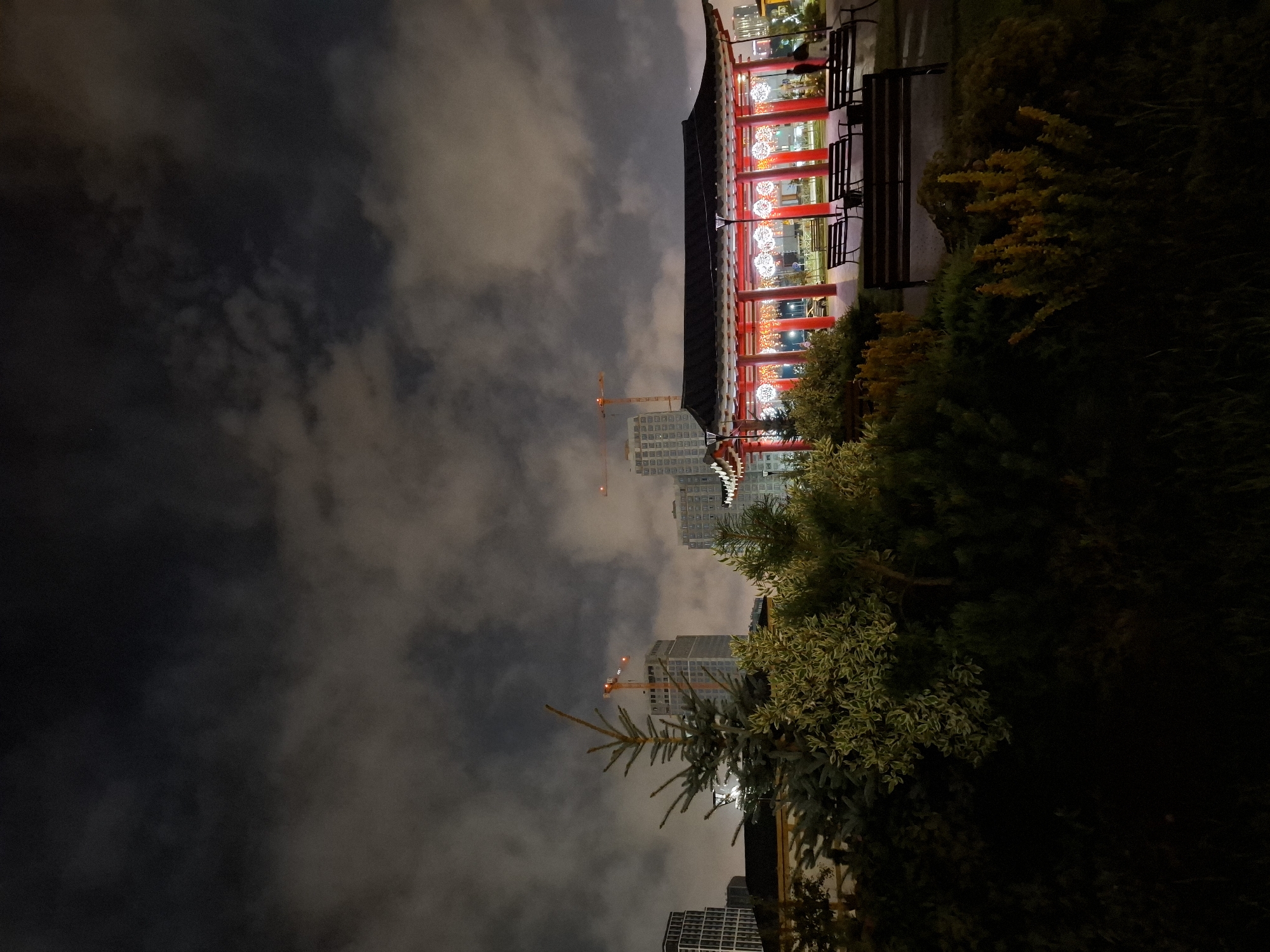 The image depicts a night scene in the city of Tyumen, Russia, featuring a traditional-style pavilion with red pillars and a dark roof, illuminated by decorative white lights. The pavilion is situated within a park or garden area, evidenced by lush green and yellow foliage in the foreground and several dark wooden benches placed in front of the structure. In the background, several tall modern buildings and construction cranes are visible against a cloudy night sky, suggesting an urban environment undergoing development. The lighting in the scene is primarily artificial, casting a warm glow on the pavilion and its surroundings while the rest of the area remains in shadow.